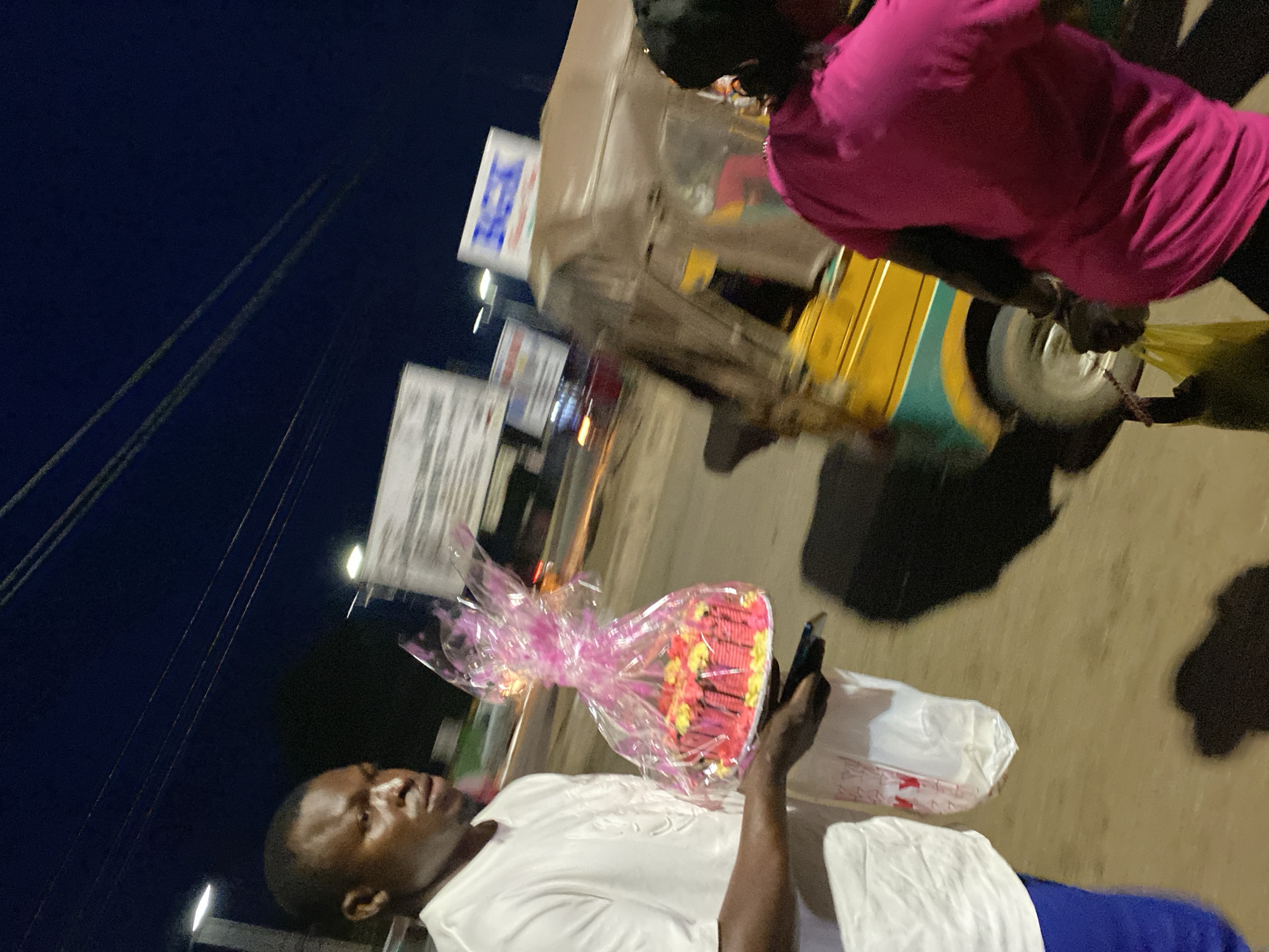 A man, wearing a white t-shirt and blue pants, walks in the foreground, holding a cake wrapped in pink plastic. He is looking to his left, with his right hand supporting the cake from underneath, and his left hand holding a dark object, possibly a phone, near his hip. Behind him, a woman in a pink shirt walks away from the camera. A yellow and green auto-rickshaw is visible to the right, with its side covered by a translucent material. Signs are visible in the background, including one that appears to say "BGK." The scene is dimly lit, suggesting it is evening or night, with artificial lights illuminating parts of the environment. The ground is paved and appears to be a street. The image is taken at an angle, and there is motion blur, indicating movement of the subjects or the camera. The overall setting suggests an urban environment in Bukuru, Nigeria.