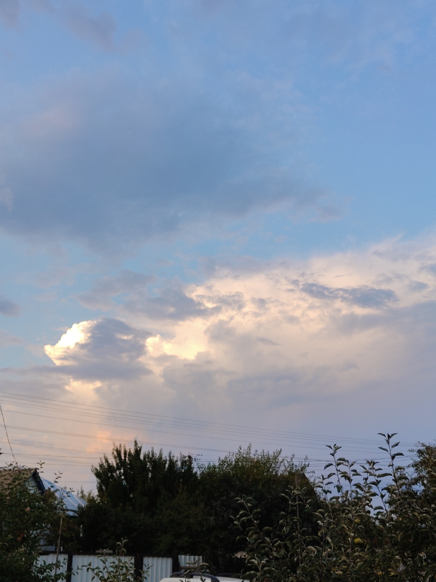 The image depicts a cloudy sky over a residential area. The sky is a mix of light blue and grey clouds, with some patches illuminated by sunlight. Below the sky, there is a dense line of trees and some houses. Power lines are visible spanning across the scene. In the foreground, there is a white fence and the branches of trees, one with a visible apple. The scene is likely a suburban or rural setting. The location is identified as Cherkasy, Ukraine.