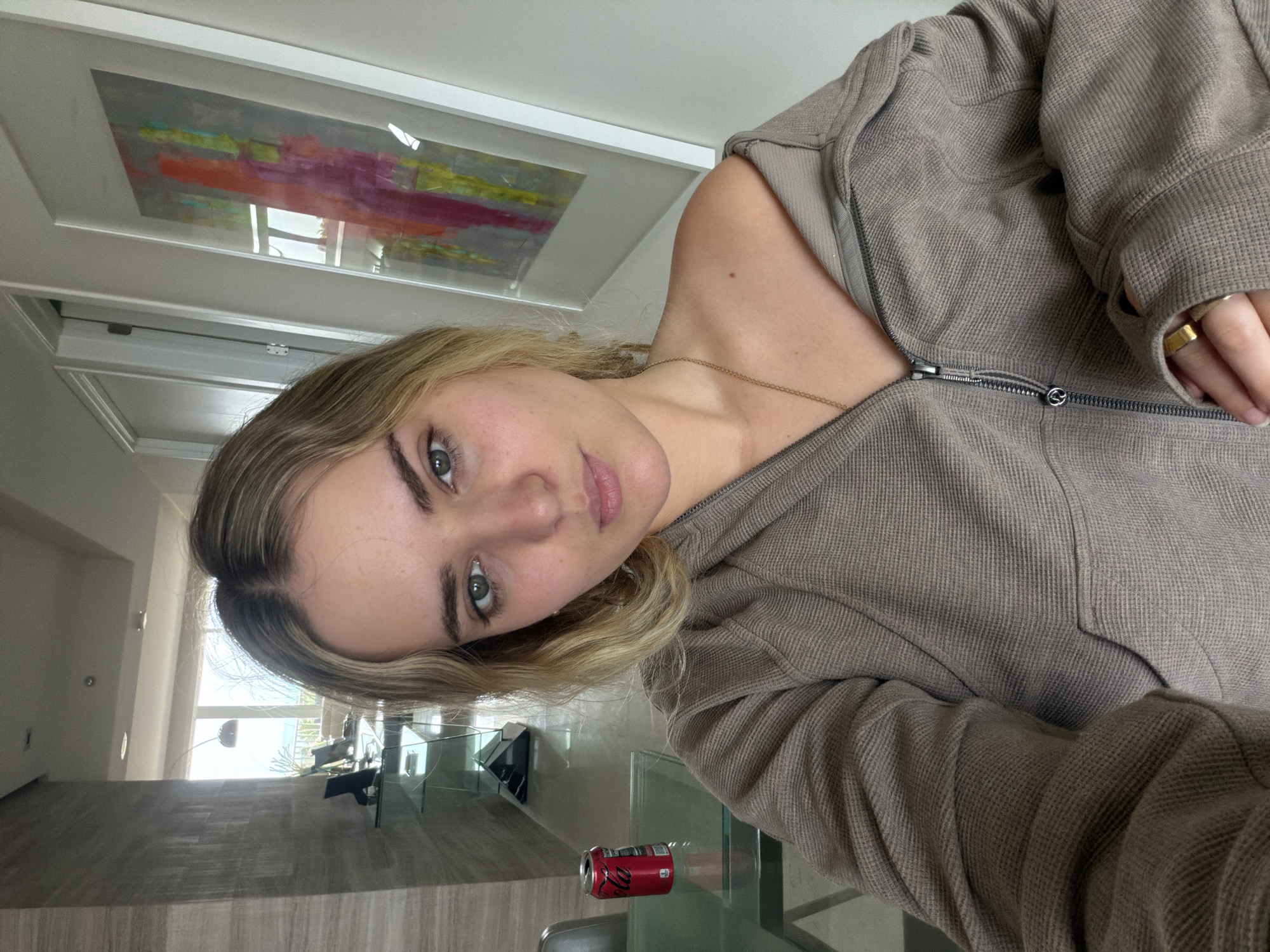 A fair-skinned individual with blonde hair and green eyes is facing the camera while wearing a beige hooded zip-up jacket and a gold ring. A gold necklace is visible. The subject is indoors, in a room with modern decor, including a glass table on which there is a crushed Coca-Cola can. A framed painting hangs on the wall. Outside the window, trees and blue water are visible. The location is likely Key Biscayne, United States of America.