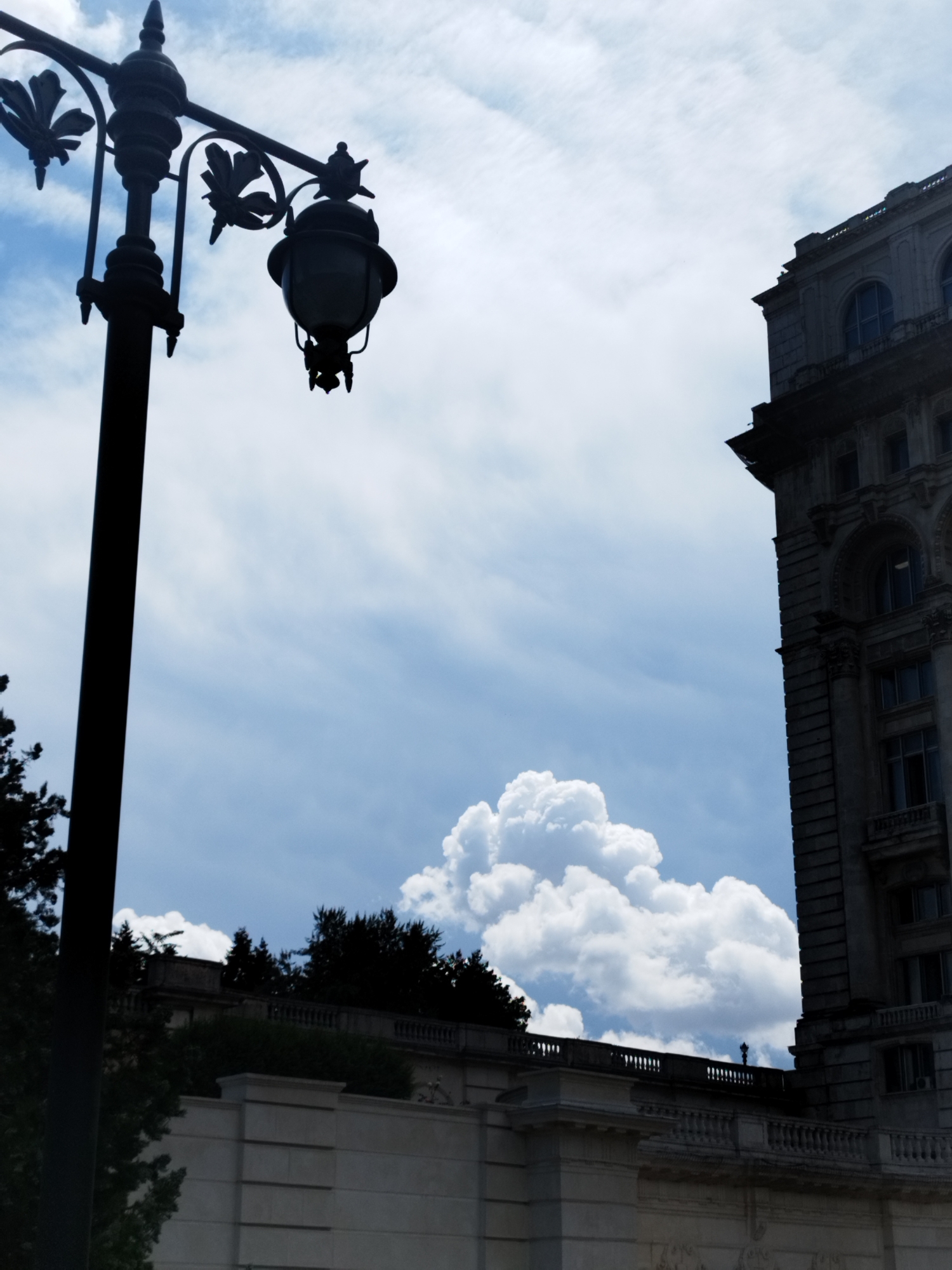 The image displays a streetlamp silhouetted against a blue sky with white clouds, partially obscuring a grand, ornate building. The streetlamp is antique, with decorative elements and a glass lantern. To the right, a tall, stone building with arched windows and intricate architectural details is visible. Below, a white stone wall with a decorative railing and trees can be seen, suggesting a park or a grand avenue. The overall impression is of an urban environment with historical architecture, possibly in a city like Bucharest, Romania, known for its neoclassical buildings.