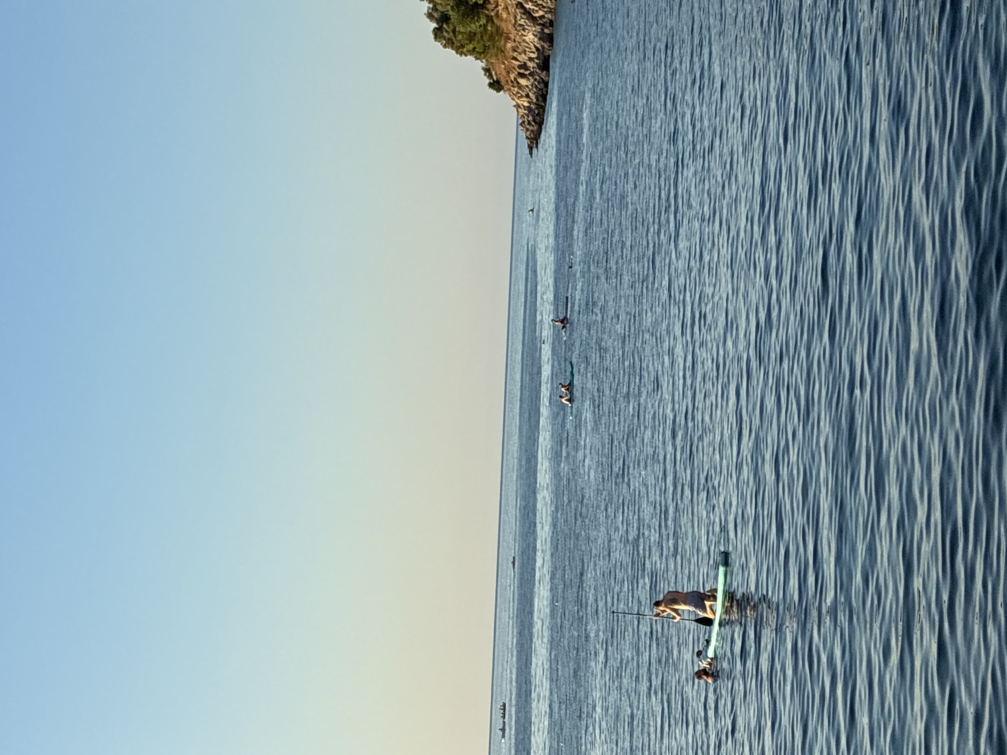 This image captures a serene ocean scene with several individuals engaged in water activities near the coast of Alanya, Turkey. In the foreground, a person is kneeling on a stand-up paddleboard, holding a paddle, with another person partially submerged in the water beside the board. Further out in the water, other individuals are visible on paddleboards or swimming. In the distance, several small boats can be seen on the horizon. The background features a rocky coastline dotted with trees, indicative of the Mediterranean landscape. The sky is clear and blue, transitioning to a lighter hue near the horizon. The water is calm, with gentle ripples reflecting the clear sky.