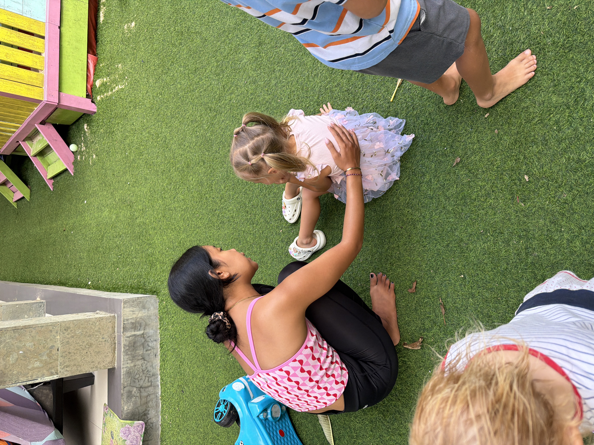 The image depicts an outdoor scene on a bright day, possibly in Kuta, Indonesia. A young woman with dark hair pulled back is crouching on vibrant green artificial turf, wearing a pink geometric-patterned tank top and black leggings. She is interacting with a young girl who is sitting on the turf. The girl has blonde hair styled in pigtails and wears a light pink dress with a sheer, floral-patterned overlay. The woman has her hand gently placed on the girl's back. Nearby, a blue toy ride-on vehicle is visible. To the right of the frame, the legs of another person, likely a child, are visible, wearing grey shorts and a striped shirt. In the background, part of a colorful structure, potentially a playhouse or playground equipment, can be seen. The environment appears to be a recreational or play area, indicated by the artificial turf and play equipment.
