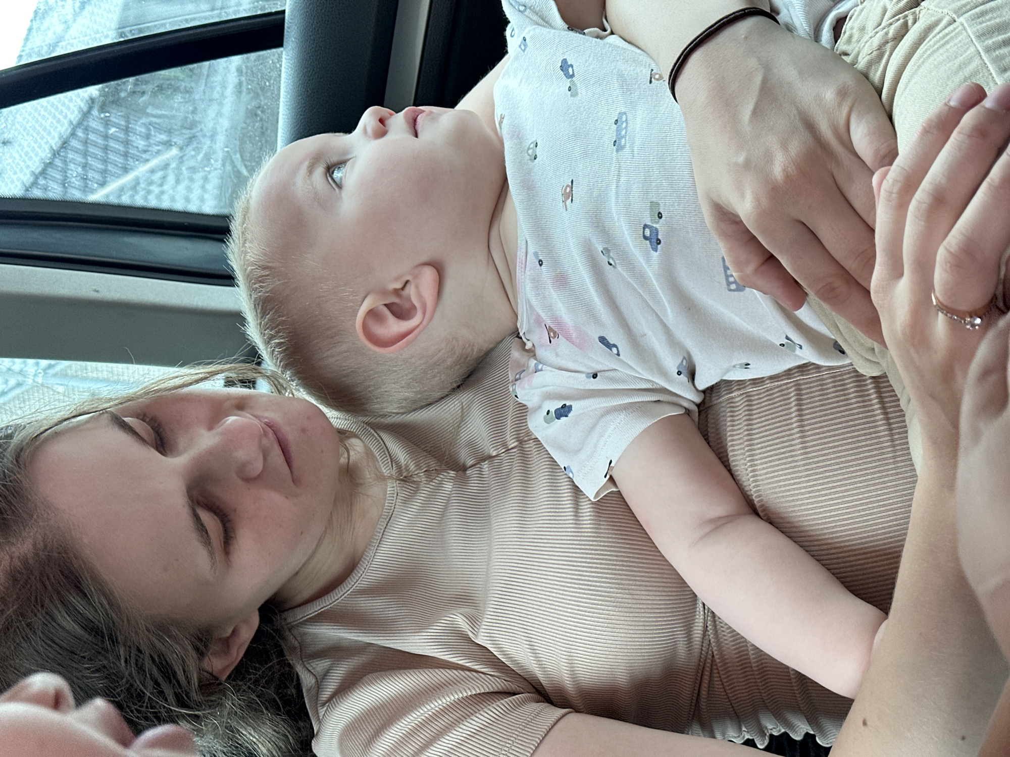 A woman in a beige ribbed t-shirt is holding a baby in her lap in a car. The woman is looking down at the baby with her eyes closed and a slight smile on her face. The baby is looking out the window with a curious expression. The baby is wearing a white onesie with a pattern of toy cars. The interior of the car is visible, including the car door and window. The background outside the window appears to be a rural landscape with some buildings. It is difficult to determine the exact location in Kutenholz, Germany from the image alone.