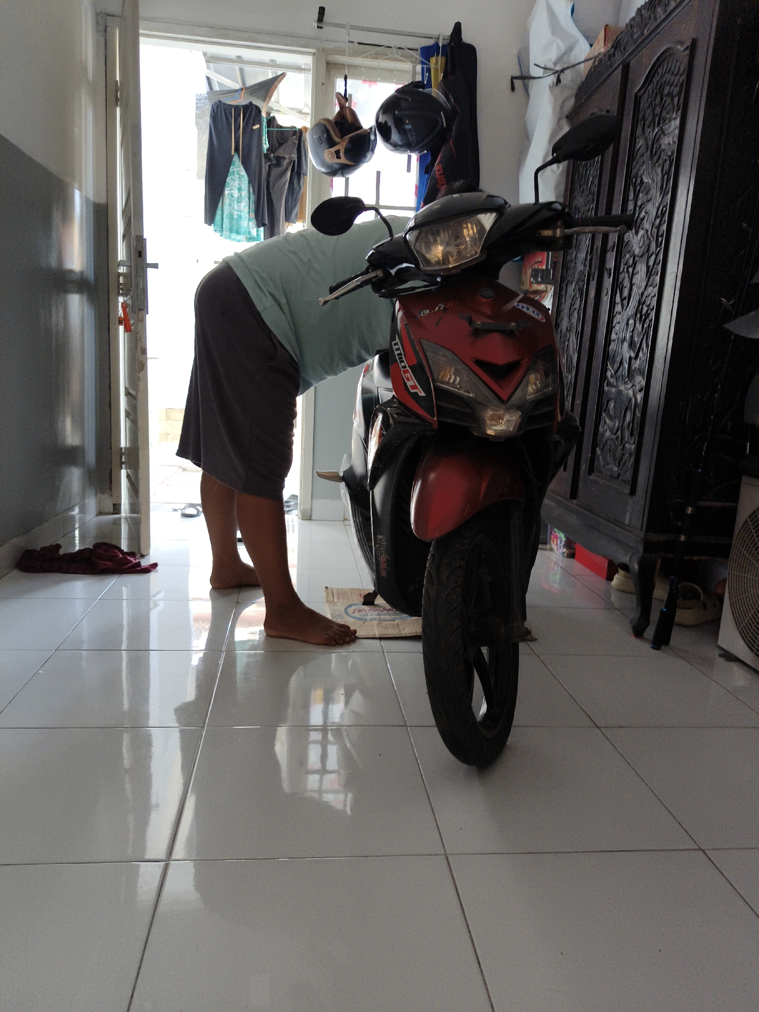 The image shows a red and black motorcycle parked indoors, possibly in a garage or a sheltered area. A person, wearing a teal t-shirt and gray shorts, is bending over the front of the motorcycle, suggesting they are interacting with it, perhaps cleaning or inspecting it. The floor is tiled and appears to be clean and well-lit, reflecting the surroundings. In the background, there's a large wooden cabinet with intricate carvings, and various items are hung on the wall, including clothes and helmets, indicating a living space or storage area. The overall impression is of a domestic or personal space in Bandar Lampung, Indonesia.