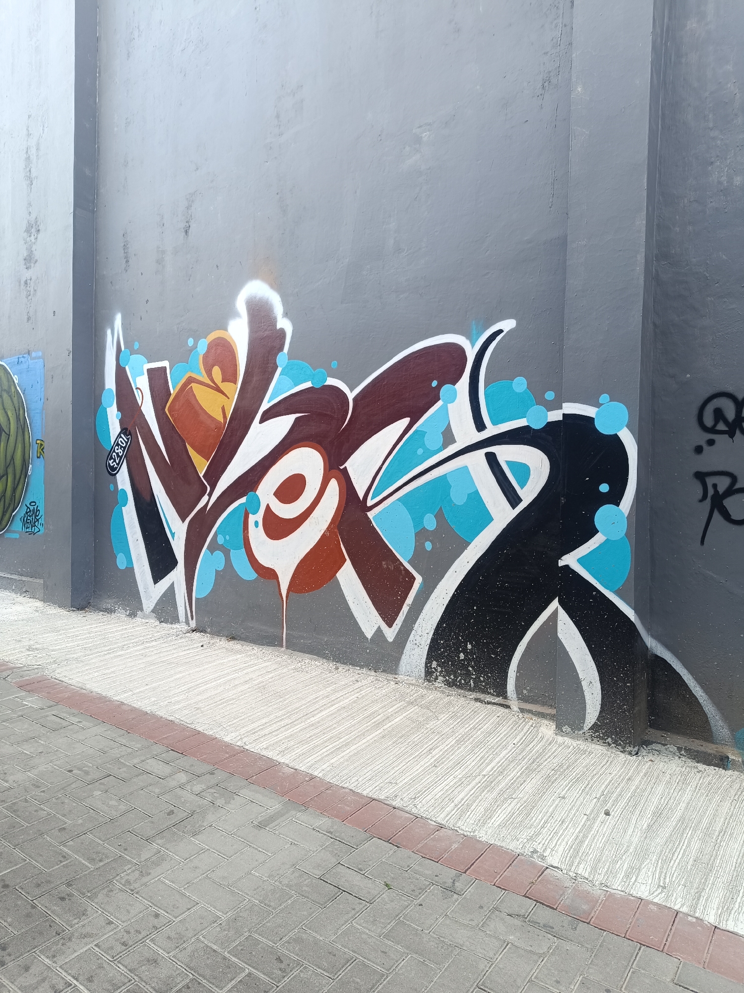 The image captures a section of a grey concrete wall adorned with vibrant graffiti, located on a sidewalk in Yogyakarta, Indonesia. The graffiti features a stylized representation of the word "WER" in bold, blocky letters, with a color palette dominated by brown, white, black, and bright blue accents forming bubbly shapes. The lettering is complex and dynamic, with sharp angles and flowing lines. To the left of the main graffiti, a partial view of another artwork featuring a green, textured sphere is visible. The foreground shows a paved sidewalk constructed with grey interlocking bricks arranged in a herringbone pattern, bordered by a strip of reddish-brown bricks. The wall itself appears weathered in places, with some minor smudges and drips of paint visible on its surface and the adjacent ground. The overall scene suggests an urban environment where street art is a prominent feature.