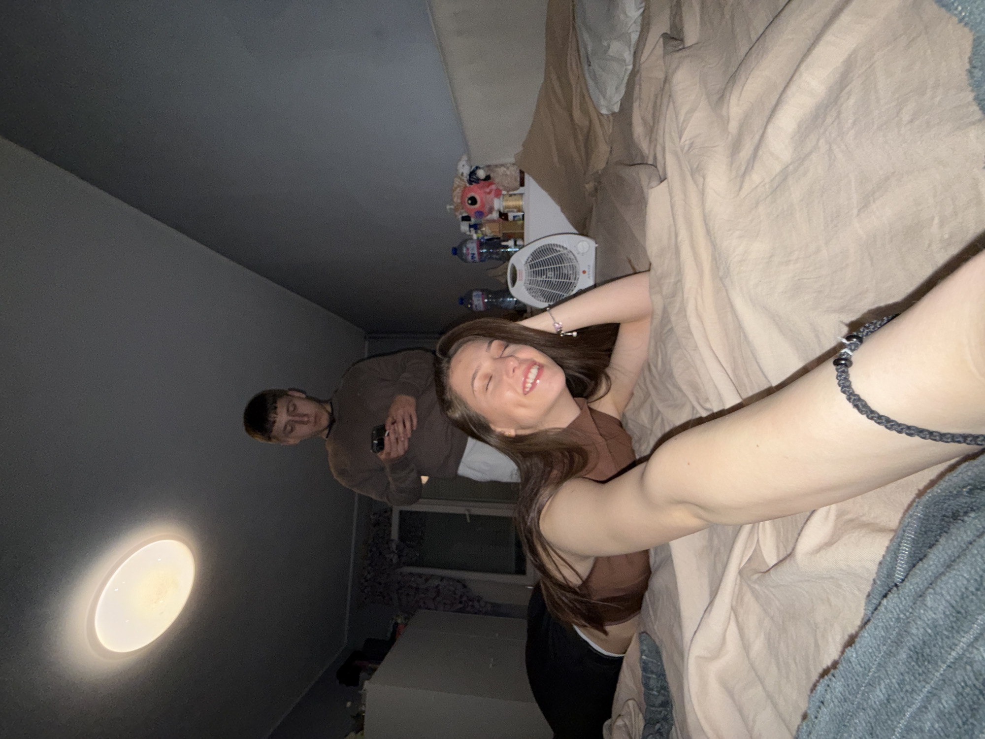 The image captures an indoor scene, likely a bedroom, from a tilted perspective. A young woman dominates the foreground, lying on a bed covered with light brown bedding. She has long brown hair, is wearing a brown halter-neck top and black pants, and is smiling broadly at the camera, seemingly taking a selfie. A dark braided bracelet is visible on her wrist.
In the background, a young man stands on the left side of the frame. He has short, dark hair and is wearing a brown long-sleeved top. He is looking down intently at a smartphone he holds in his hands. Above him, a bright, circular light fixture illuminates the dark grey ceiling.
To the woman's left, a shelf or nightstand is visible, cluttered with various items. These include a small white fan with the brand "AVCO" written on it, several plastic water bottles, and a collection of stuffed animals, such as a pink pig and a brown fluffy toy. A grey textured blanket or piece of clothing is partially visible in the bottom right corner of the frame. The walls are a plain dark grey. The artificial lighting suggests the photo was taken indoors during the evening or night. The mood conveyed is relaxed and informal.