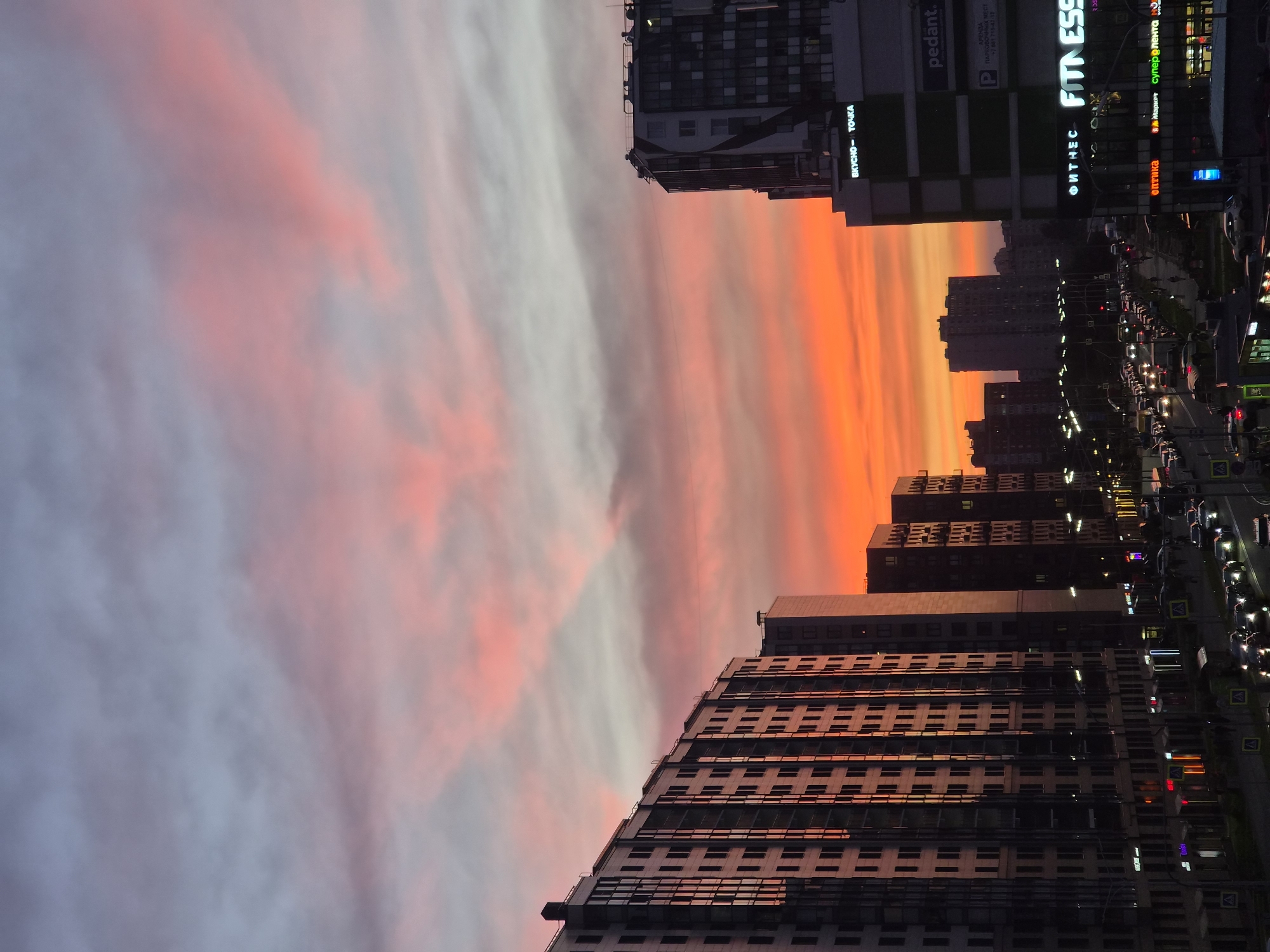 A street-level view of a city skyline at dusk in Kudrovo, Russia, is captured. The sky is illuminated by a vibrant sunset, with hues of orange, pink, and purple painting the clouds. Tall, modern residential buildings with numerous windows dominate the left side of the frame, their facades reflecting the warm colors of the sky. Across the street, more buildings rise, including a structure with a prominent "FITNESS" sign illuminated in bright white letters. Below, a busy street is lined with traffic, cars with their headlights on, and a few pedestrians are visible. The scene suggests an urban environment transitioning from day to night, with the natural beauty of the sunset contrasting with the man-made structures of the city.