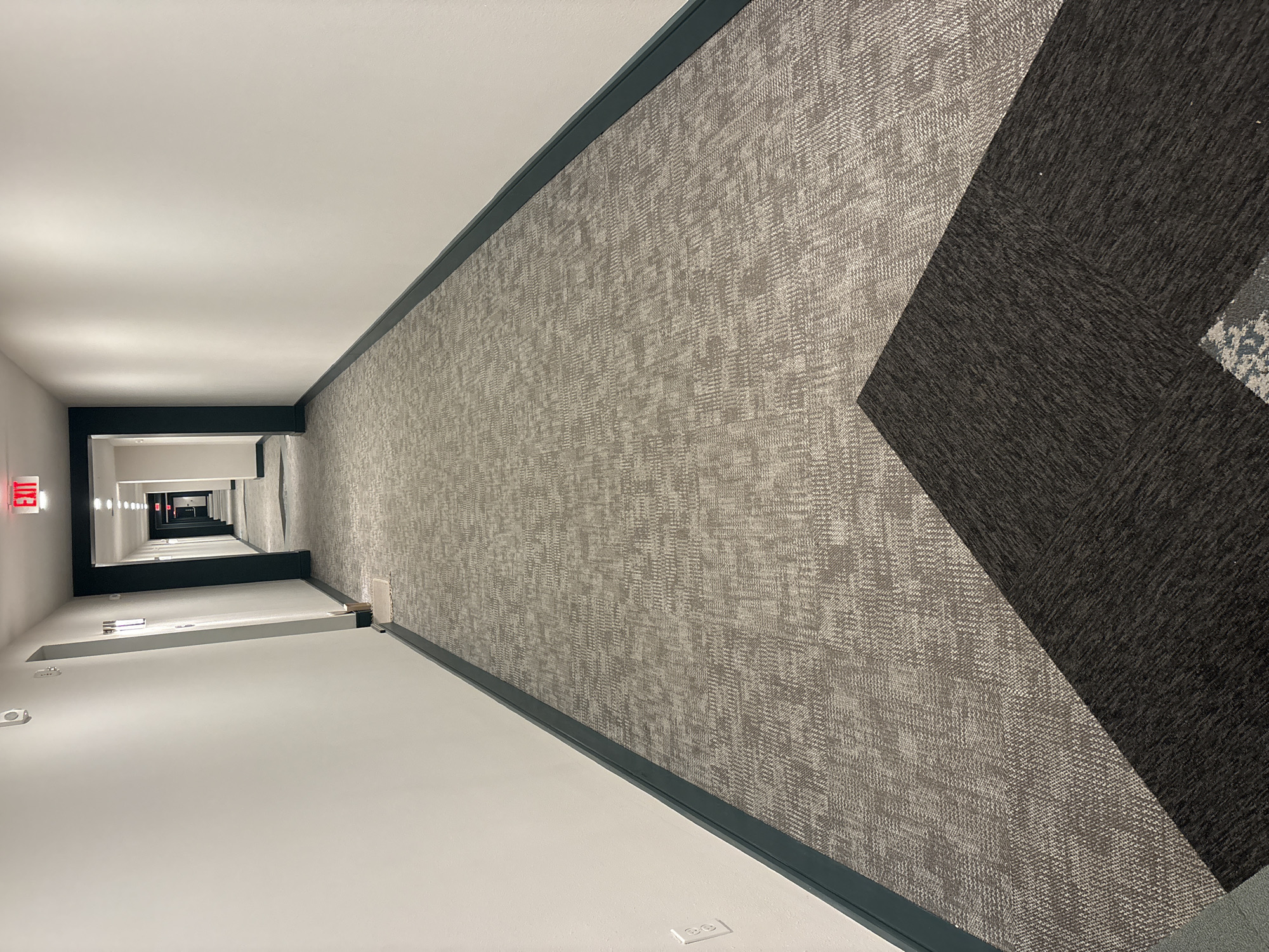 The image shows a long, empty hotel or apartment building hallway with patterned grey carpet. The walls are painted white, and there is a dark teal baseboard running along the bottom. An illuminated red "EXIT" sign is visible at the far end of the hallway. The hallway recedes into the distance, with several doorways visible on the right side. There are no people present in the image. The scene is clean and well-lit, with overhead lights contributing to the illumination. In the foreground, a section of darker carpet is laid over the lighter patterned carpet, creating a contrasting geometric pattern. A white electrical outlet is visible on the left wall. The location is specified as Lakewood Ranch, United States.