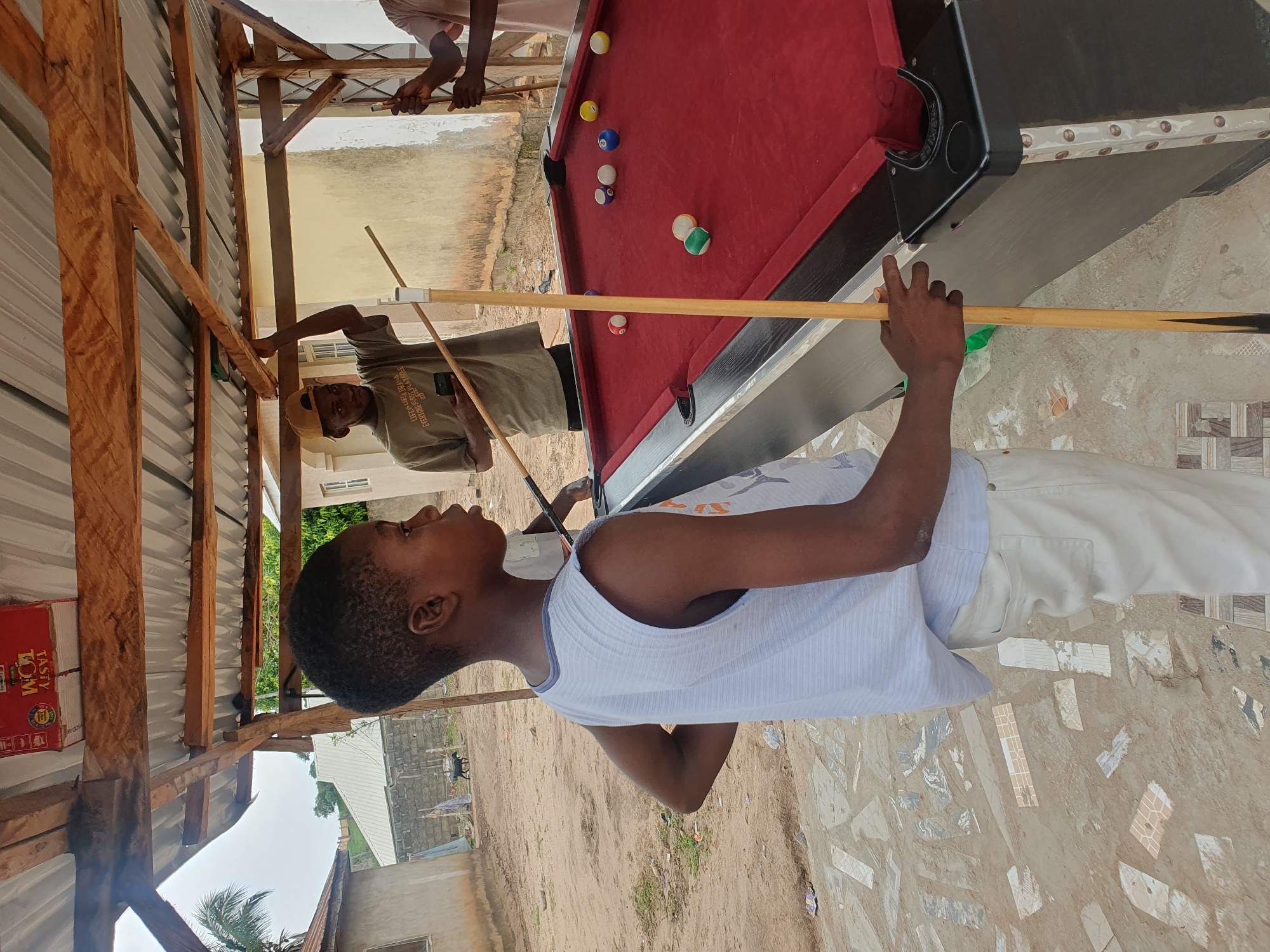 In Lokoja, Nigeria, a young boy stands by a red pool table under a makeshift wooden shelter. He wears a white tank top and off-white pants, holding a pool cue. The shelter has a corrugated metal roof supported by wooden beams. Behind him, another person in a tan t-shirt and a baseball cap plays pool, holding a phone in one hand and a cue in the other. A third person, partially visible, also holds a pool cue. The ground is a mix of sand, dirt, and scattered broken tiles. In the background, simple buildings and a small black goat are visible. The sky is overcast.