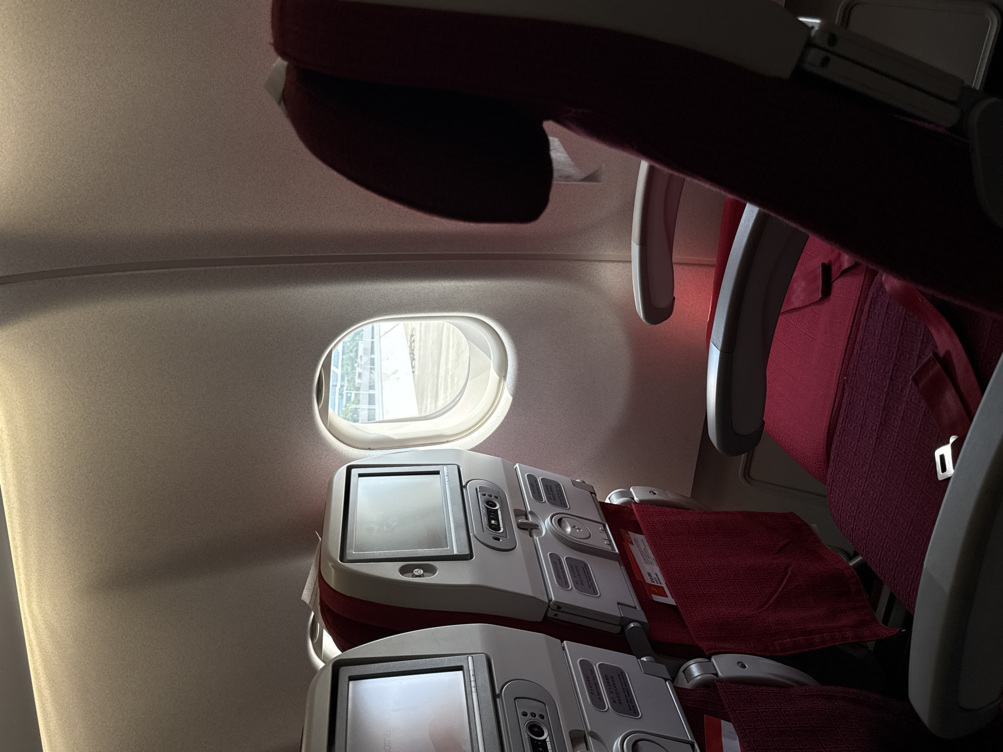This is a photograph taken inside an airplane cabin, likely at Xiaoshan District, China, depicting a row of seats by the window. The seats are upholstered in a deep red fabric with a repeating geometric pattern. In the foreground, a seat with a headrest is visible, along with its armrests and seatbelt. To the left, two more seats are partially visible, each equipped with an entertainment screen and a seat pocket holding magazines or safety cards. The window, oval-shaped, offers a view of an outdoor area that appears to be an airport tarmac, with sunlight filtering through. The interior of the cabin is lit by overhead lights, casting shadows that highlight the texture of the seats and the curved walls of the aircraft.