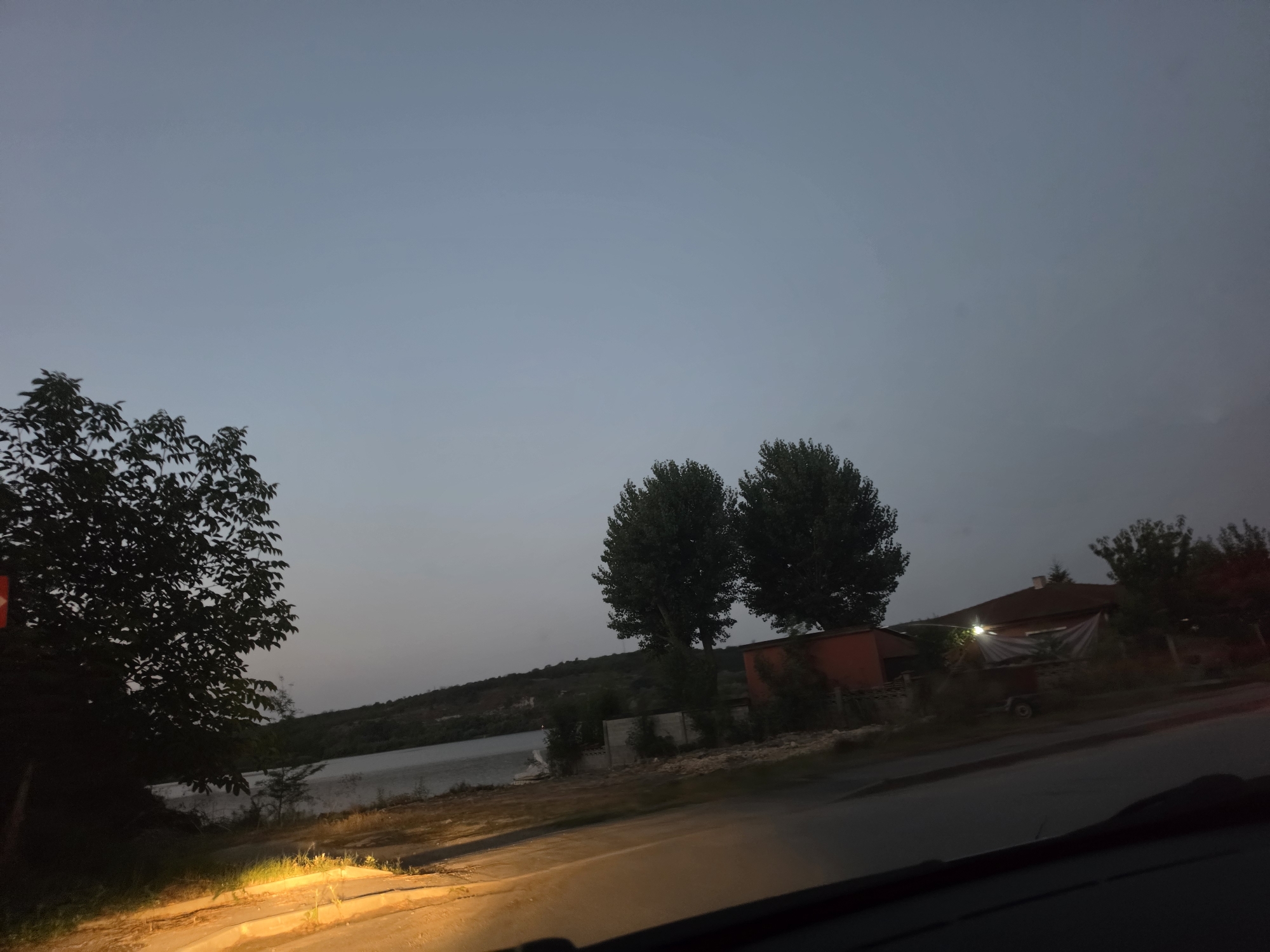 The image depicts a twilight scene with a body of water on the left, bordered by a tree-lined hill in the background. In the foreground, a road curves towards the right, with artificial lighting illuminating a section of it. On the right side of the image, several structures are visible, including a building with a reddish-brown roof and illuminated windows, a smaller orange structure, and a trailer. Two tall trees with full canopies stand between these structures and the water. The overall atmosphere is dim, characteristic of dusk or early dawn. The location is identified as Strashimirovo, Bulgaria.