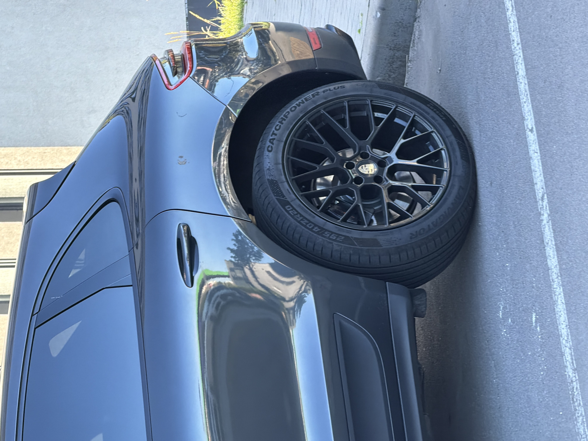 A dark grey Porsche Macan is parked on a paved surface, possibly a street or parking lot, in front of a modern building with light-colored walls. The car's rear passenger side is prominently featured, showing the alloy wheel, tire, fender, and a portion of the door and rear quarter panel. The tire has the brand name "CATCHPOWER PLUS" visible. The environment appears to be a daytime scene, with soft shadows indicating indirect sunlight. There are no people visible in the image. The location is identified as Kyiv, Ukraine.