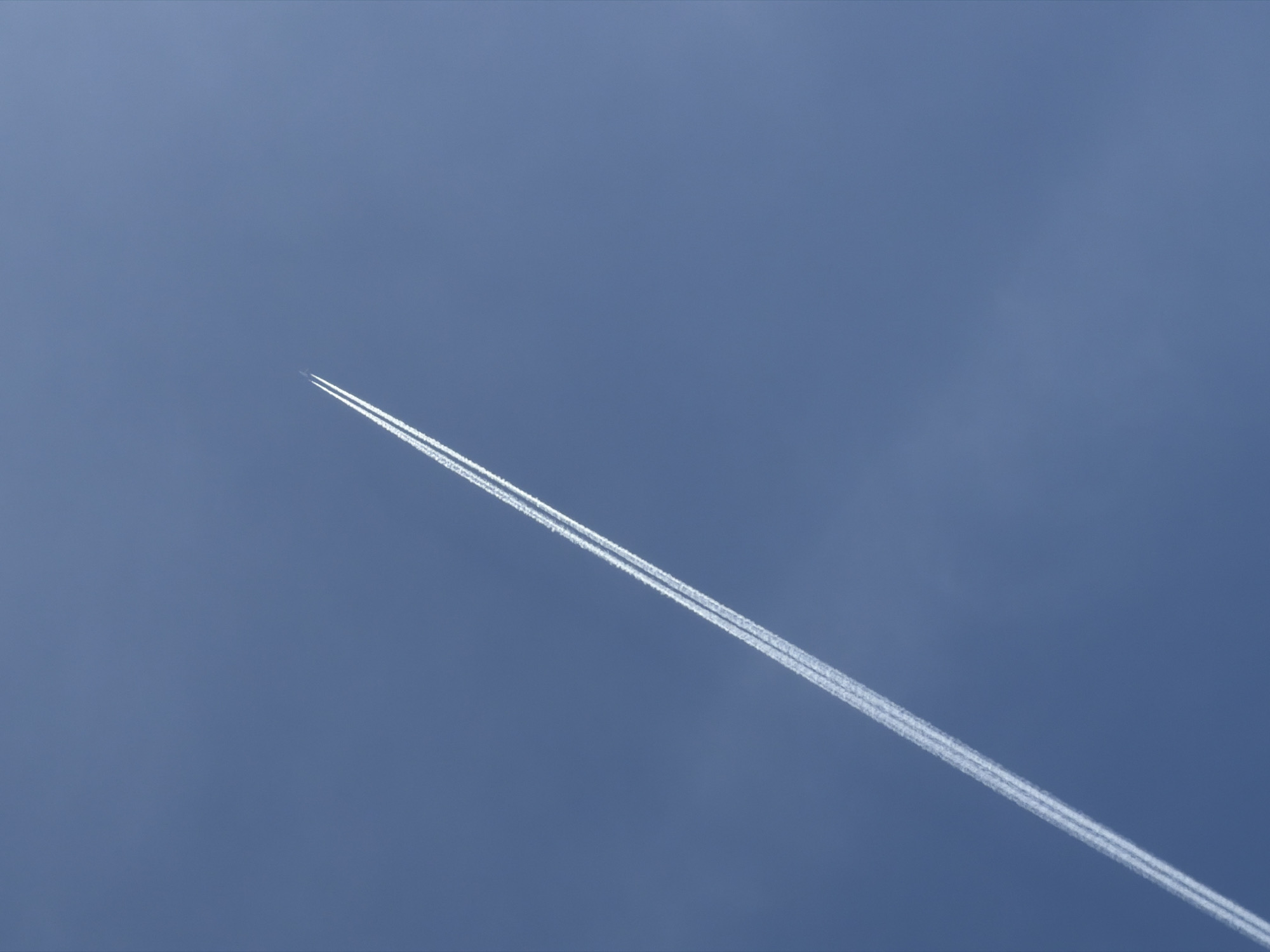 The image shows a contrail in a blue sky. The contrail is a white line stretching diagonally across the frame from the lower left to the upper right. The contrail appears to be produced by a jet plane, which is not visible. The sky is a solid blue color with slight variations in tone. There are no other objects or people visible in the image. The image is not in the city of Ol'khovo, Russia.