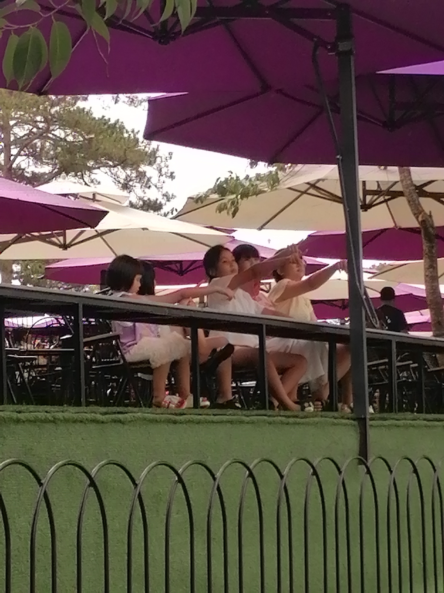 Four people are sitting at an outdoor table. The person on the right is pointing. The three to the left have their arms outstretched. There are tables and black chairs in front of and to the left of the seated people. There is a black railing in the foreground. The patio area is covered with purple and white umbrellas. Trees are visible. The scene appears to be in Da Lat, Vietnam.