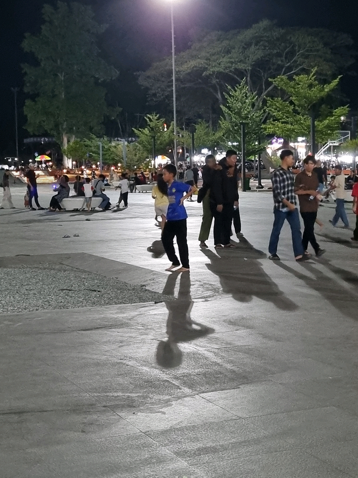 The image depicts an outdoor public space at night, likely Tanamodindi, Indonesia, characterized by paved ground, trees, and artificial lighting. Several individuals are present, engaged in various activities. In the foreground, a young person in a blue shirt and black pants walks with their shadow cast on the ground. To their left, a group of people are seated on a bench; one person in a striped shirt is holding a blue object. Further back, a group of men stand together, some with their arms around each other. Other individuals are scattered throughout the scene, some walking, others standing. In the background, trees and more people are visible, along with what appears to be a food stall with colorful umbrellas and decorative lights. The overall atmosphere suggests a lively evening gathering in a community space.
