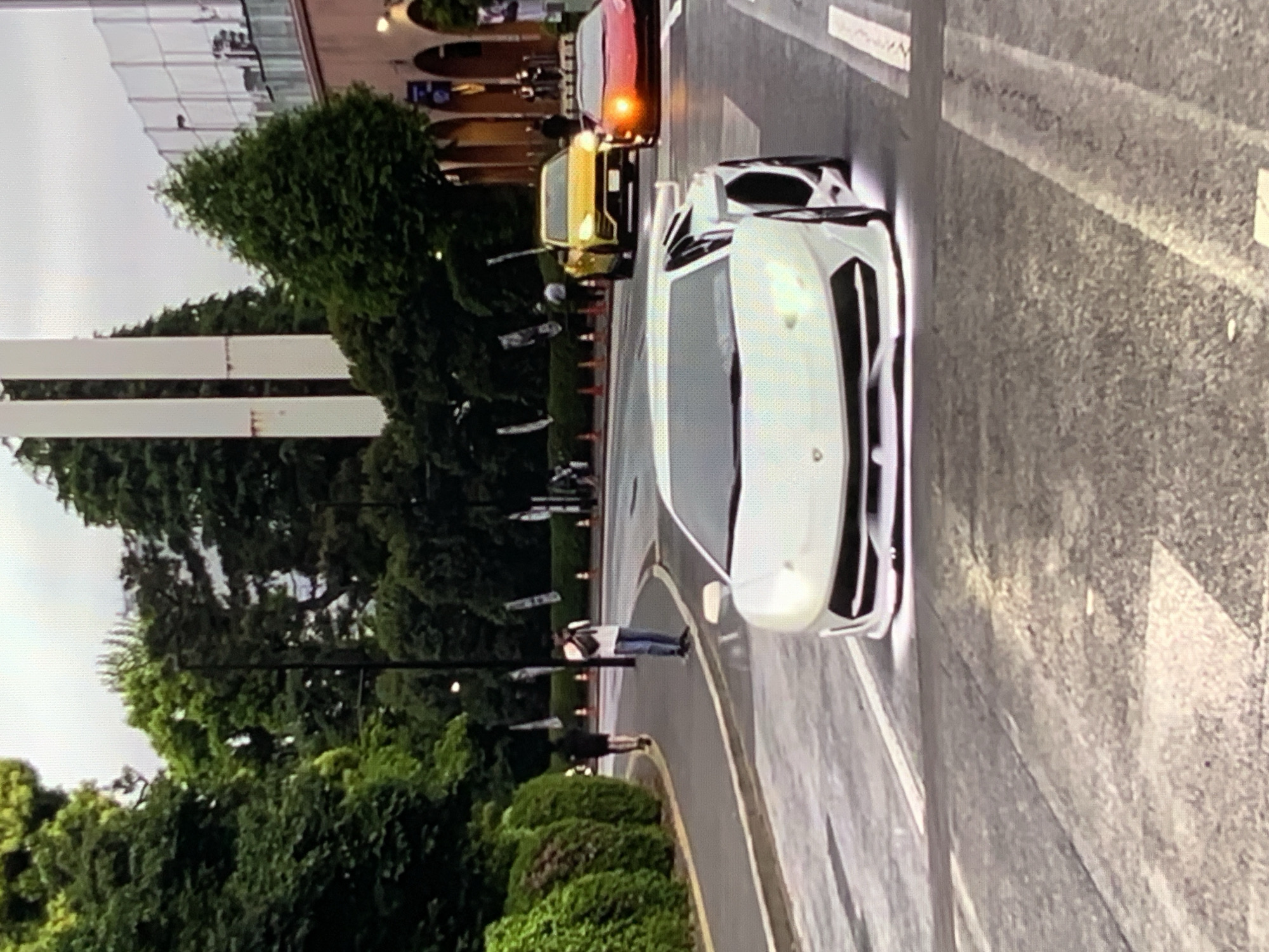 In an urban environment, possibly a city street with lush green foliage lining the side, a white Lamborghini is prominently featured in the foreground, angled towards the right of the frame. The car has its headlights illuminated, casting a bright glow. Further down the road, a bright yellow SUV is visible, followed by a red sports car with its brake lights on.
Two individuals are present on the sidewalk to the left. One person, dressed in dark clothing, is walking away from the camera. Another individual, wearing a white t-shirt and blue jeans, is standing and appears to be taking a photo with a phone. They are positioned on a paved pathway adjacent to a manicured hedge.
The background reveals a mix of tall, slender white structures and dense green trees, suggesting a park-like or landscaped area within the city. Beyond the trees, a building with an arched facade and a fence are discernible, hinting at a sports facility or a similar establishment. The sky is overcast, indicating a diffused light condition. The road surface shows markings and some wet patches, suggesting recent rain or a damp environment.