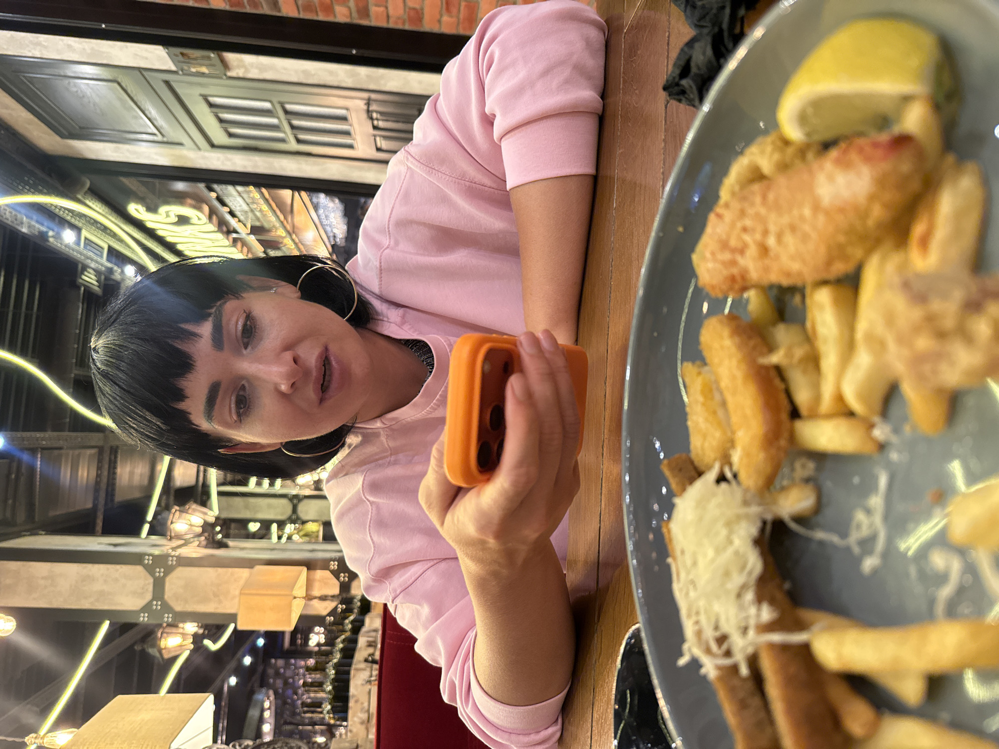 An over-the-shoulder medium shot captures a young woman with short black hair and bangs, wearing a pink sweatshirt, sitting at a wooden table. She is looking down at her orange smartphone, which she holds in her hands. Her mouth is slightly open, as if she is speaking or about to speak. Large hoop earrings adorn her ears. In the foreground, a plate of food is visible, featuring what appears to be fish and chips, with a wedge of lemon on the side. The background suggests an indoor setting, possibly a restaurant, with ambient lighting and decorative elements on the walls. The overall impression is casual, with the woman engaged with her phone while at a meal.