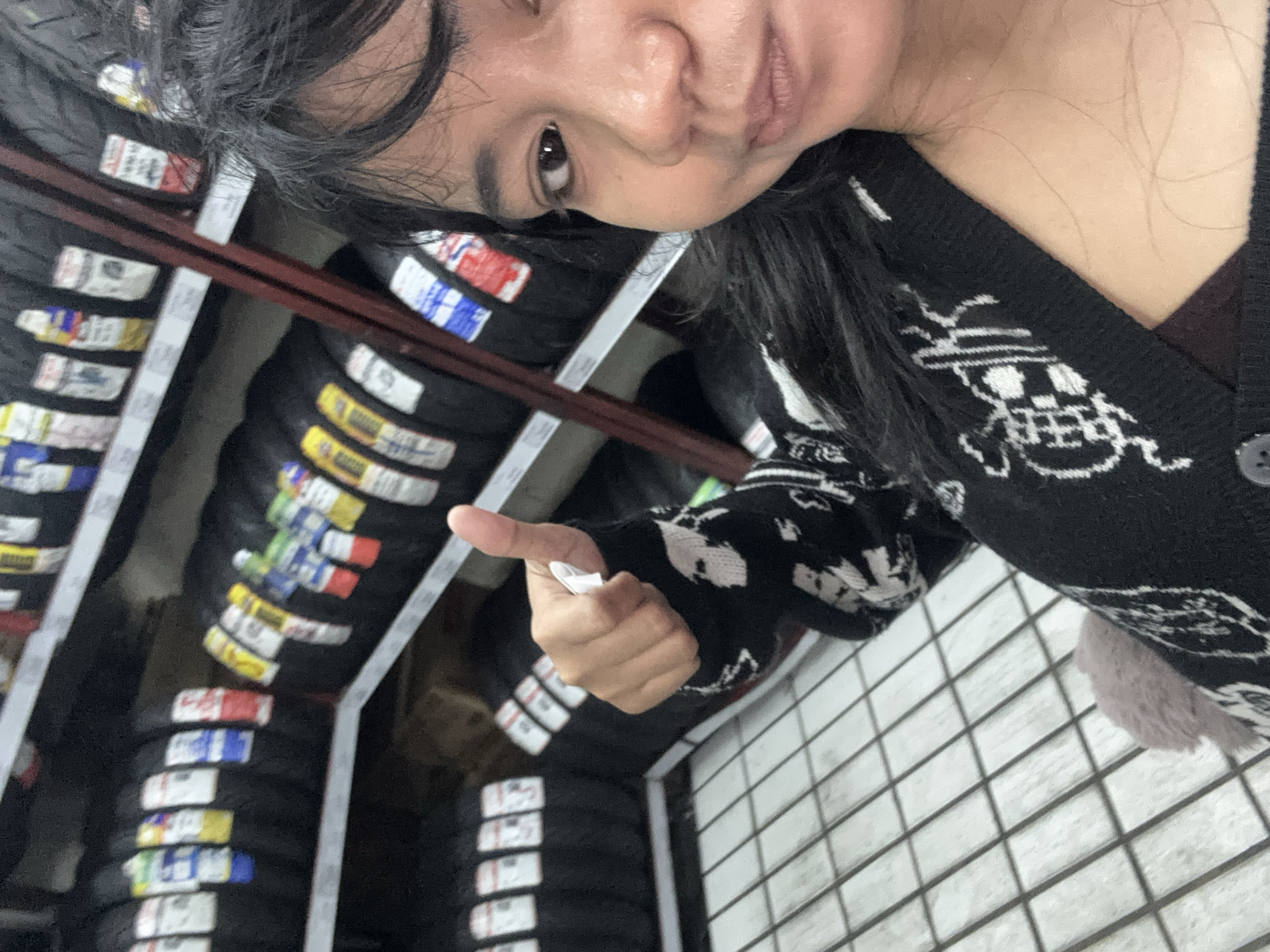 The image shows a person in a store, possibly a tire shop, given the rows of tires visible in the background on shelves. The person is taking a selfie, with their face partially visible in the foreground, and their thumb extended upwards in a "thumbs up" gesture. They are wearing a black and white patterned sweater. The environment appears to be an indoor retail space with tiled flooring. Without additional information or context specific to North Jakarta, Indonesia, the precise location within the city cannot be determined.