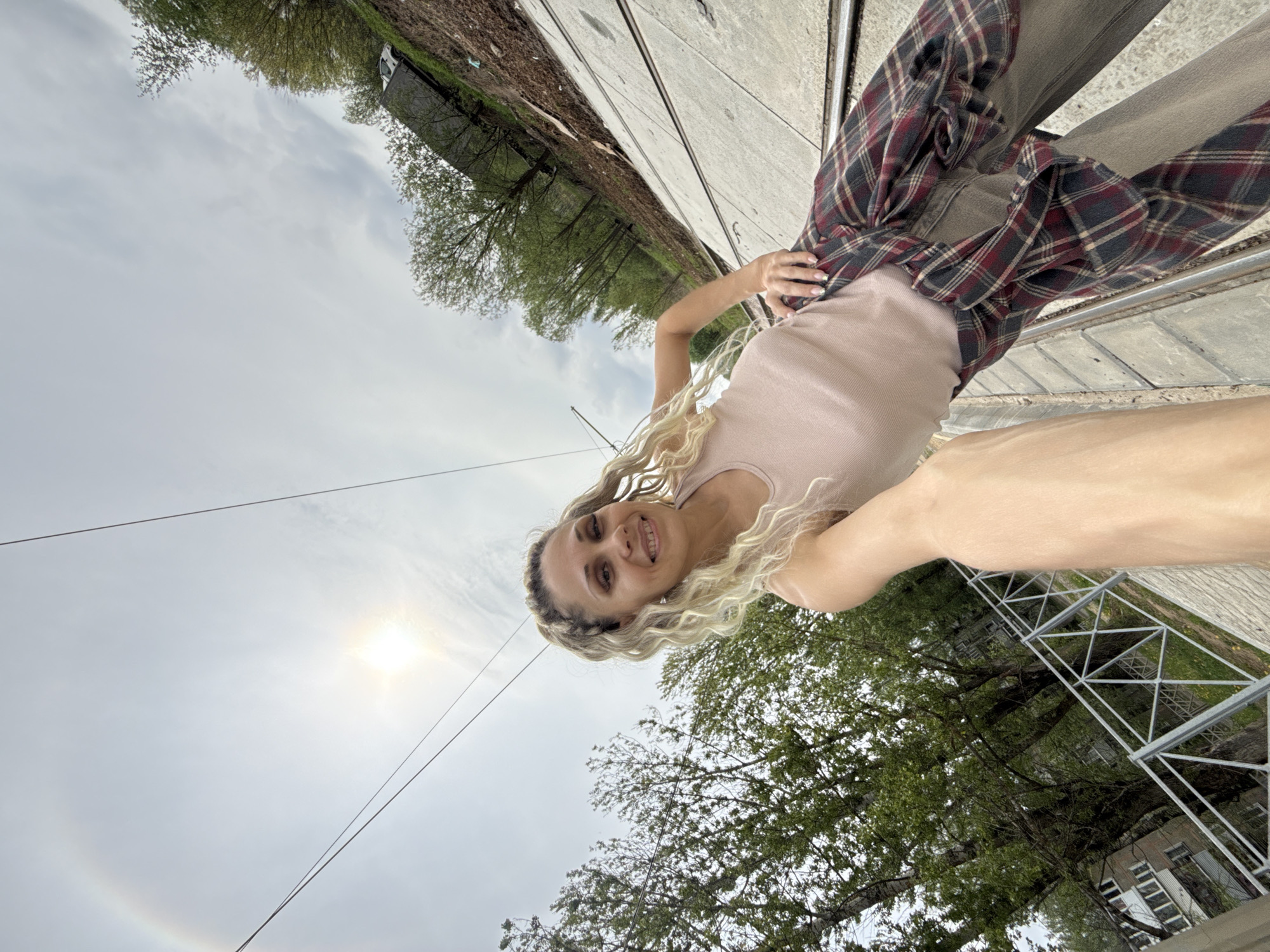 A medium-shot selfie angled upward from below features a woman standing on a sidewalk in Rusanovo, Russia. She is positioned approximately in the center of the frame, smiling directly at the camera. She has blonde hair in a long, wavy style. She wears a light pink tank top, a plaid shirt tied around her waist, and gray jeans.
Behind her, the sidewalk runs along the top of the frame, with some vegetation and a brick building visible in the background. Below her is a large tree with green foliage and a metal structure of a tower with geometric patterns and an apartment complex. The sky is visible above the tree and tower, showing a diffused light with a rainbow colored ring around the sun. Power lines run diagonally across the sky. The image is taken during daylight hours under somewhat cloudy conditions.