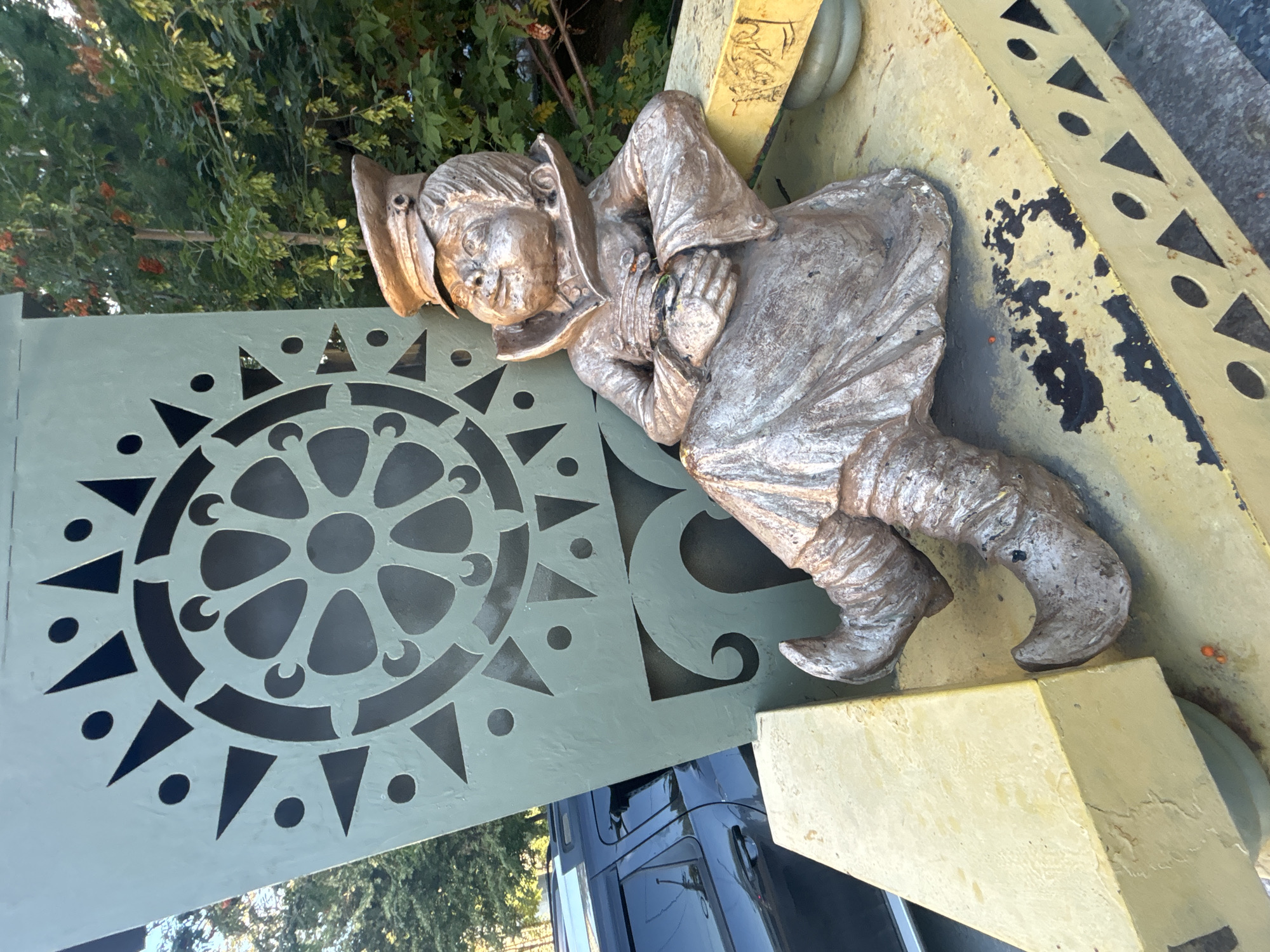 A bronze sculpture of a gnome in a cap and uniform is seated on a yellow bench. The gnome is leaning back and appears to be resting. Behind the gnome is a tall, green, triangular structure with a circular, decorative cutout. The scene is outdoors, with trees and foliage in the background. A dark-colored car is partially visible to the left of the sculpture. This sculpture is located in Novosibirsk, Russia, specifically at the Novosibirsk Zoo.