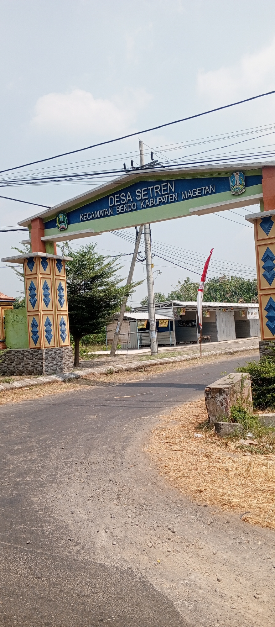 This is a full-frame, eye-level, outdoor, daytime shot of the entrance to Desa Setren, Kecamatan Bendo, Kabupaten Magetan, Indonesia. The archway, a prominent feature, is decorated with the Indonesian flag, the national emblem, and colorful patterns. Text on the archway reads "DESA SETREN KECAMATAN BENDO KABUPATEN MAGETAN". A paved road leads towards the archway, flanked by dry grass and some vegetation. In the background, there are a few buildings, possibly shops or market stalls, and power lines crisscross the sky. A single Indonesian flag is visible on a flagpole to the right of the archway. The sky is mostly clear with a few scattered clouds.