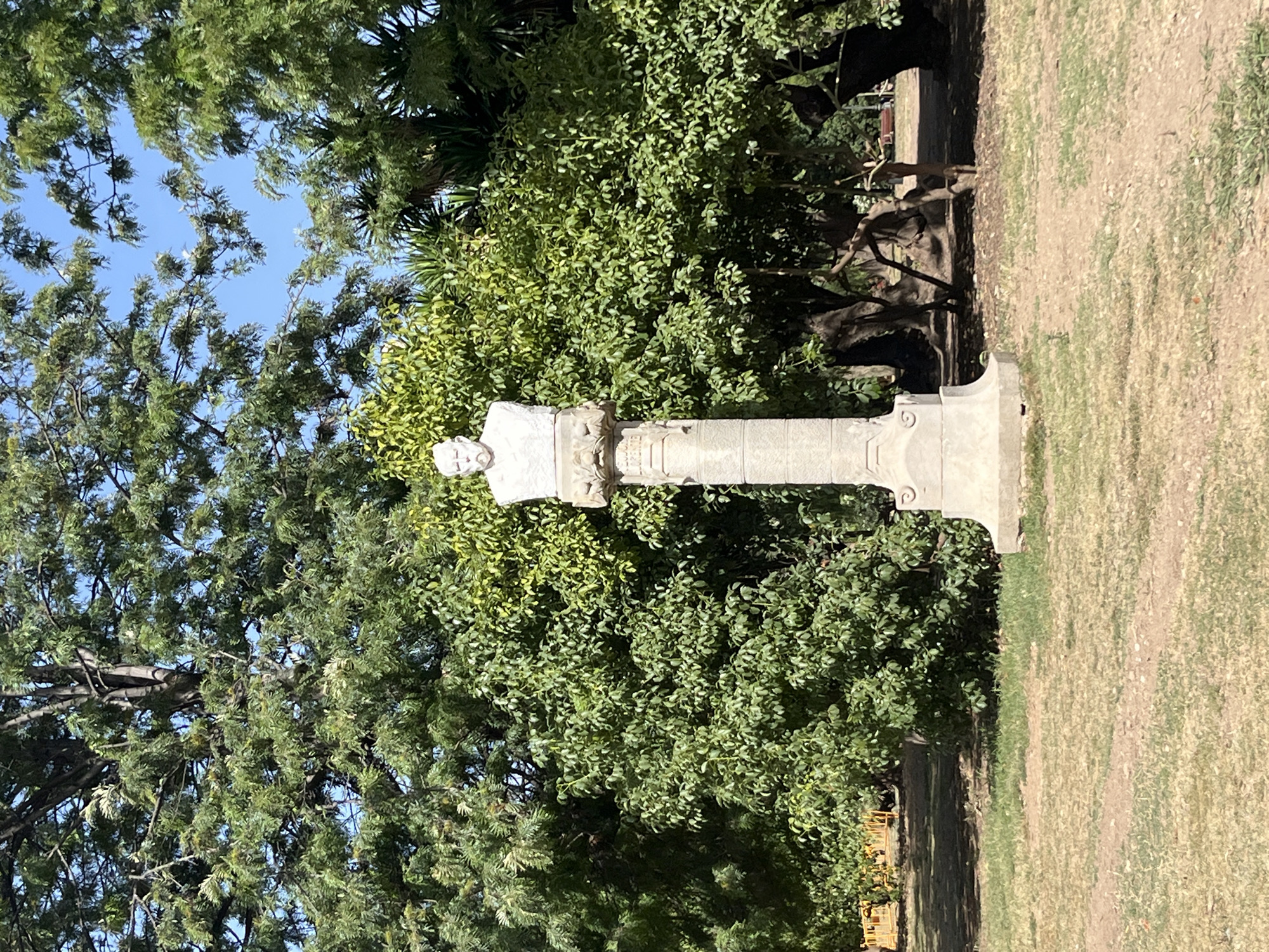 A white marble bust of a man with a mustache and beard, wearing a jacket, stands atop a tall, fluted stone pedestal. The pedestal is adorned with decorative carvings at the top. The monument is situated outdoors in a park-like setting, with lush green trees and a clear blue sky in the background. The ground in front of the monument is covered with dry grass and dirt. The overall impression is one of a public monument in a tranquil, verdant space. This monument is located in Parc de la Ciutadella in Barcelona, Spain, dedicated to Pompeu Fabra, a linguist and philologist.