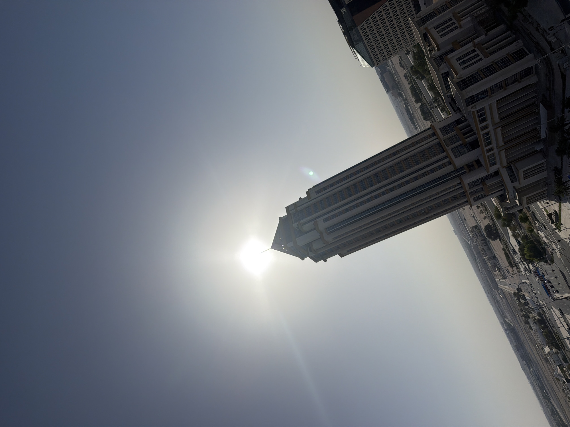 A high-angle, slightly rotated outdoor photograph captures a prominent, multi-story building with a light-toned facade, a multi-tiered base, and a sharply pointed spire. The sun is positioned directly behind the spire, creating intense backlighting, a prominent starburst, and lens flares that extend across the upper left quadrant of the image. The sky transitions from a clear, darker blue-grey at the top left to a hazy, bright white around the solar disk. To the right, a second, wider building with a dark upper section and a light-colored facade featuring a repeating geometric pattern is partially visible. Below the central building, a multi-lane road network with visible vehicles traverses the foreground. The background reveals a vast, flat landscape, appearing arid or undeveloped, dotted with sparse low-rise structures, some trees (possibly palms), and distant infrastructure, characteristic of the city of Unknown, Unknown. The scene is brightly illuminated by direct daylight; no specific individuals are discernible.