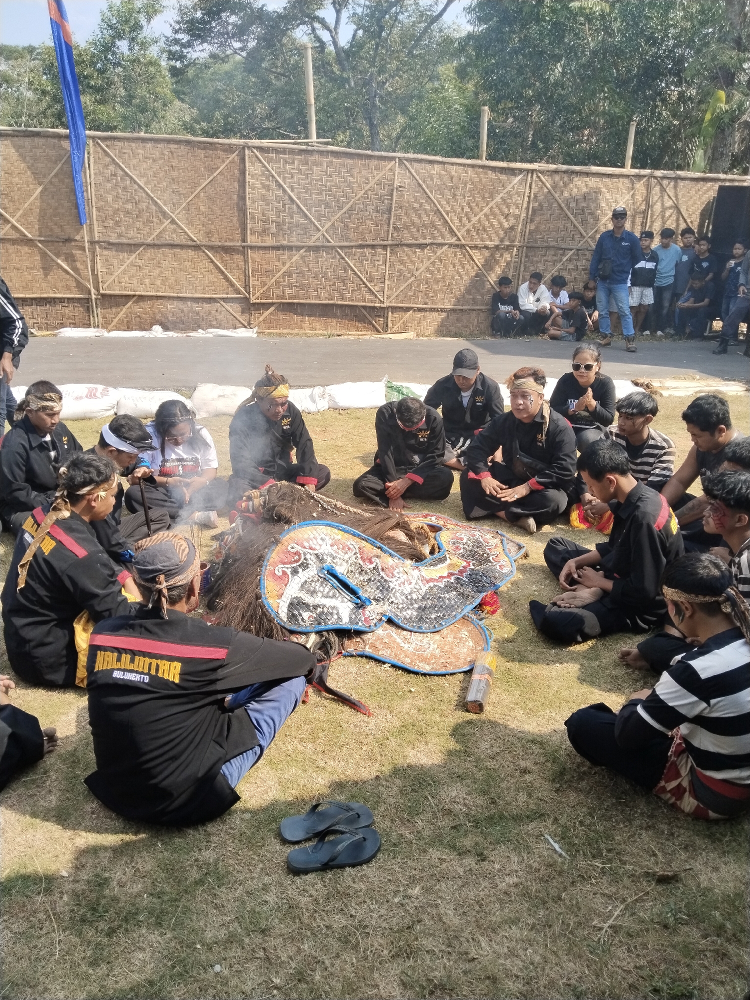 In Mojogedang, Indonesia, a group of people are gathered outdoors on a dry, grassy field in front of a bamboo wall. A portion of a blue and orange flag is visible on the left side. The people, mostly young men, are seated in a circle around a large, ornate object that appears to be part of a costume or prop. Smoke rises from the center of the circle, suggesting a ritual or ceremony is taking place. Some individuals are wearing matching black uniforms with red and yellow accents, while others are dressed in casual clothing. The overall atmosphere appears to be one of communal gathering and cultural observance.