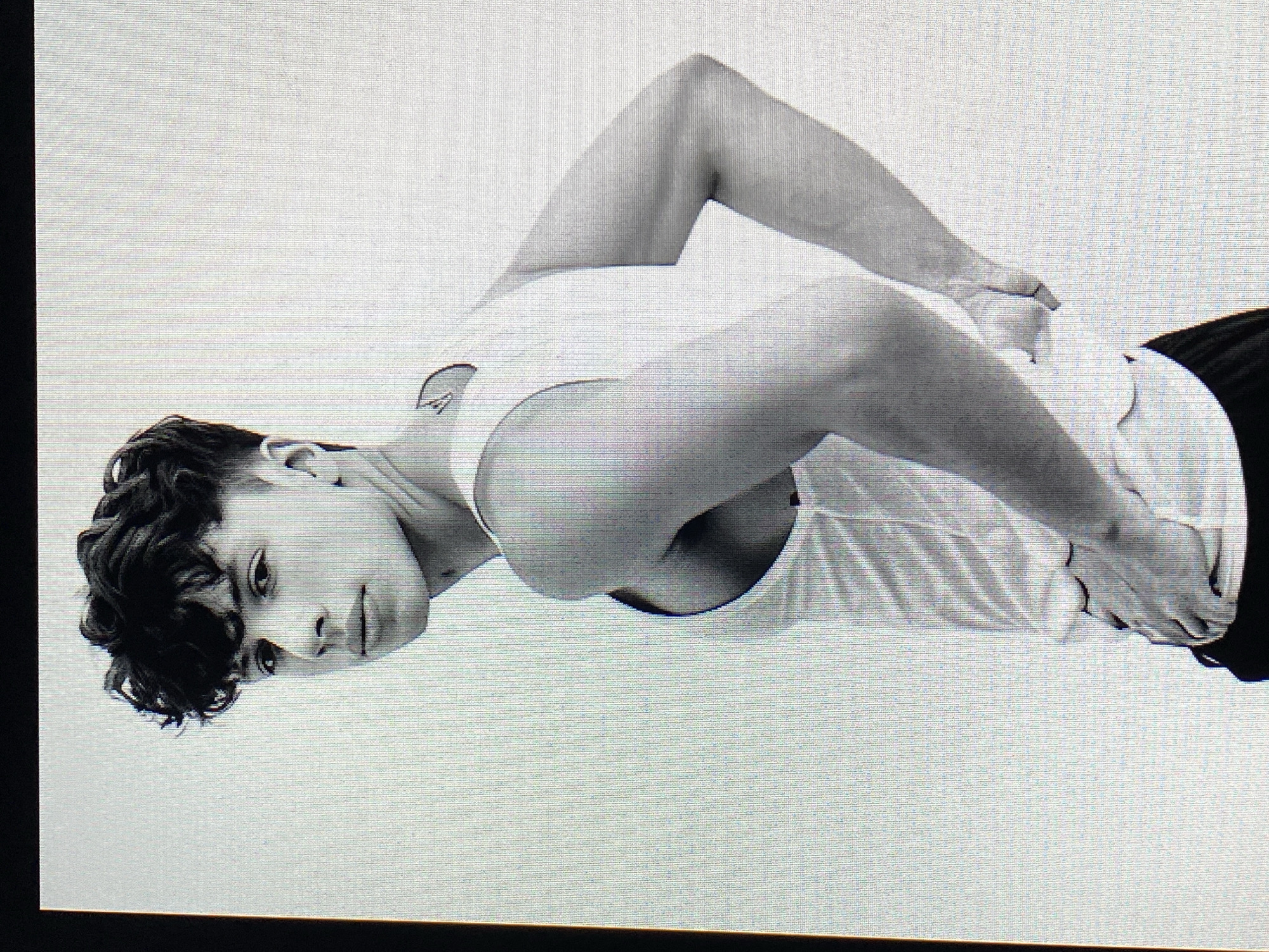 A black and white, medium shot photograph captures a young man with dark, curly hair styled in a fade haircut. He is positioned in profile, facing towards the left of the frame, with his head turned to look directly at the camera. He is wearing a white tank top and appears to be standing against a plain white background. His left hand is resting on his hip, while his right arm is bent with his hand behind his back. A small, dark tattoo is visible on the back of his neck. The lighting creates subtle shadows that highlight the contours of his face and arms. The image is a posed studio shot with no discernible city or environmental context, and therefore, its location within Poznan, Poland cannot be determined.