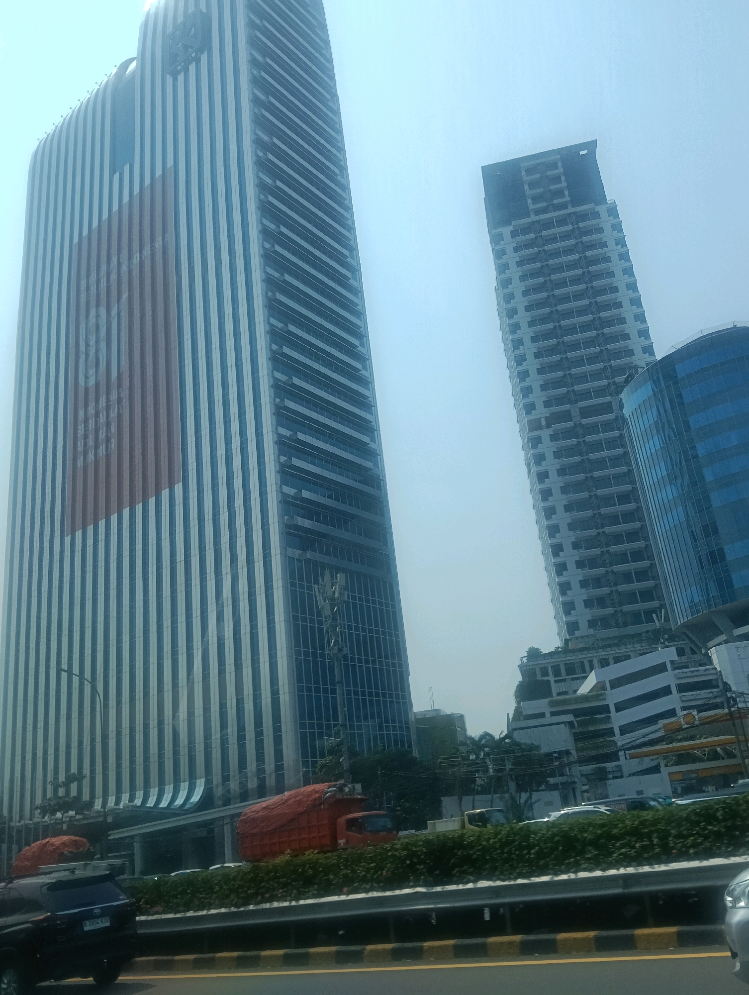 In South Jakarta, Indonesia, the image captures a dynamic urban landscape dominated by towering skyscrapers under a hazy sky. The primary focus is a colossal, modern skyscraper with a facade of vertical blue and white stripes. A large, reddish banner is prominently displayed on its upper section, featuring a stylized number "8" and text in Indonesian, which appears to be a celebratory or informational message.
To the right of this main structure, another tall building is visible, its upper floors appearing unfinished or under construction, showcasing a grid of window openings. Further to the right, a curved building with a blue glass exterior adds another dimension to the cityscape.
In the foreground, a highway or elevated road is depicted with traffic. A black SUV with license plate "B 2854 KIW" is parked or moving in the lower left quadrant. Two orange dump trucks, covered with orange tarps, are situated on the side of the road, suggesting a construction or transport activity. Green foliage lines the median of the road. The overall scene depicts a busy commercial and transportation hub within a metropolitan area.