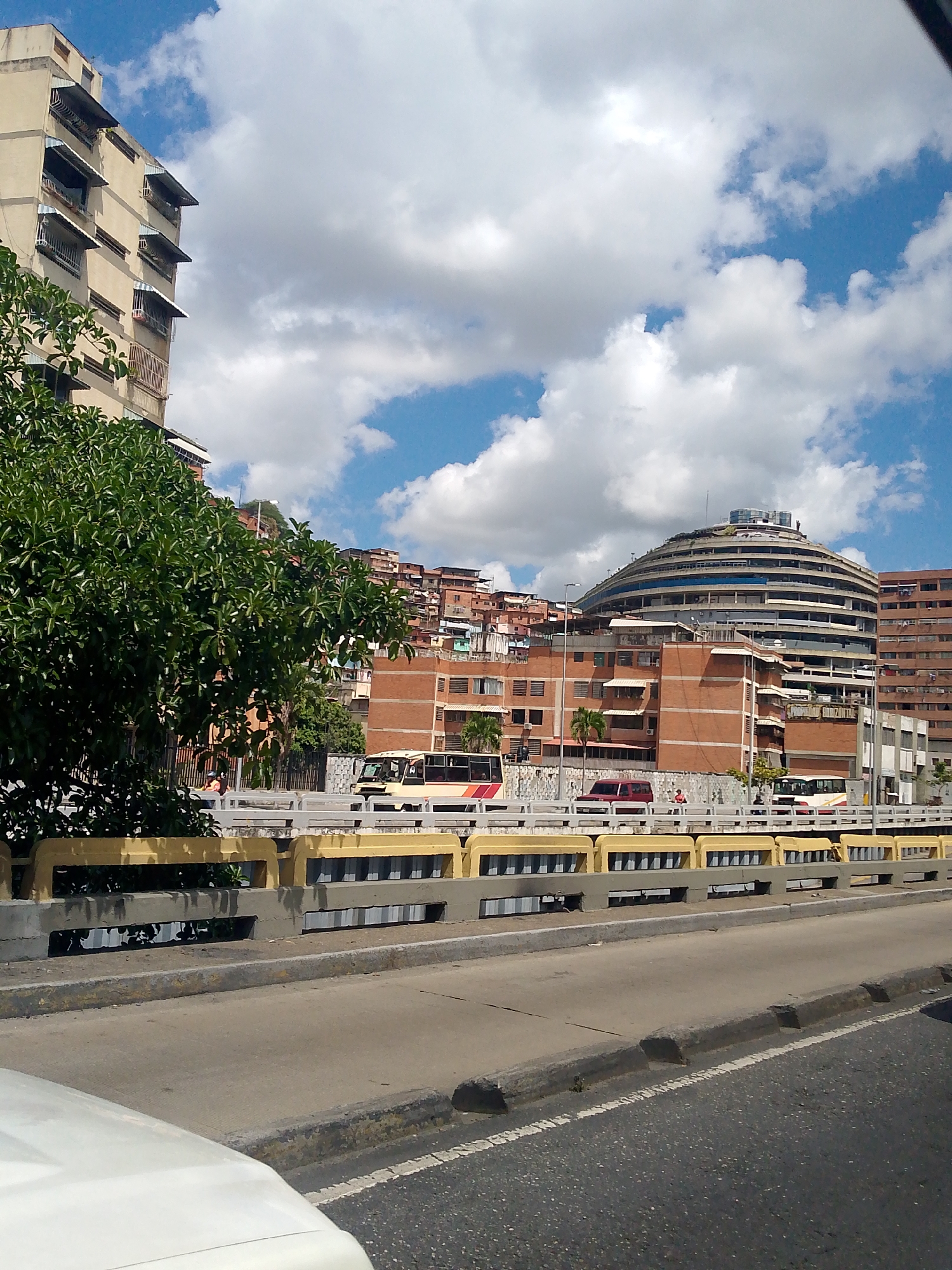 This daytime image captures a vibrant urban scene in Caracas, Venezuela, under a bright sky with scattered white cumulus clouds. The photograph is taken from inside a vehicle, with part of a white car visible in the lower left foreground.
The scene is dominated by a multi-level concrete bridge or overpass running horizontally across the midground. Its concrete railings are painted yellow and gray, with gaps revealing glimpses of the ground below. Above and behind this structure, the city unfolds.
To the right, a distinctive, large, spiraling circular building, known as El Helicoide, stands prominently, surrounded by other multi-story brick and concrete structures. These buildings appear residential and commercial, with some older structures featuring awnings over windows. On the hillsides in the background, a dense collection of smaller, more informal houses, typical of barrios or shantytowns, are visible, extending up the natural topography.
To the left, a tall, light-colored apartment building with numerous windows and awnings occupies the frame, partially obscured by a large, lush green tree in the mid-left.
Traffic is present on the bridge. Several buses are visible, along with a red van or SUV. A few people can be seen near the vehicles, appearing to be passengers or pedestrians. The road in the immediate foreground shows asphalt pavement with a white dividing line. The overall impression is that of a bustling city environment on a clear, sunny day. No discernible text is visible in the image.