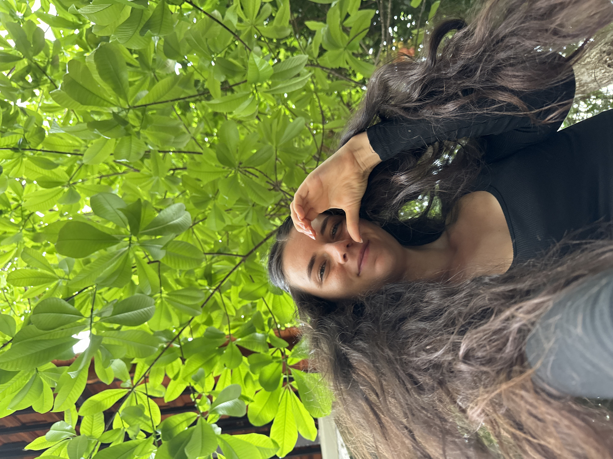 This low-angle portrait features a young woman looking directly at the camera, set against a vibrant green canopy of leaves. The woman, with long, dark, wavy hair, occupies the lower right portion of the frame. She wears a dark, ribbed long-sleeved top and has a serene expression, with a subtle smile. Her left hand is raised to her face, with fingers forming a heart shape, or possibly shielding her eyes from the bright light.
The majority of the image, particularly the upper and left sections, is filled with lush, light-green foliage. The leaves are intensely bright, suggesting strong daylight filtering through them from above, creating a backlit effect that highlights their verdant color. In the lower-left corner, beneath the dense leaves, a hint of a reddish-brown structure, possibly a roof, is visible.
The scene conveys a bright, sunny daytime setting, indicative of warm weather. The upward perspective and the woman's pose suggest a casual outdoor photo, possibly a selfie. The abundant, vibrant foliage points to a natural, possibly tropical or subtropical environment, aligning with the location context of Mũi Né, Vietnam. There is no visible text in the image.