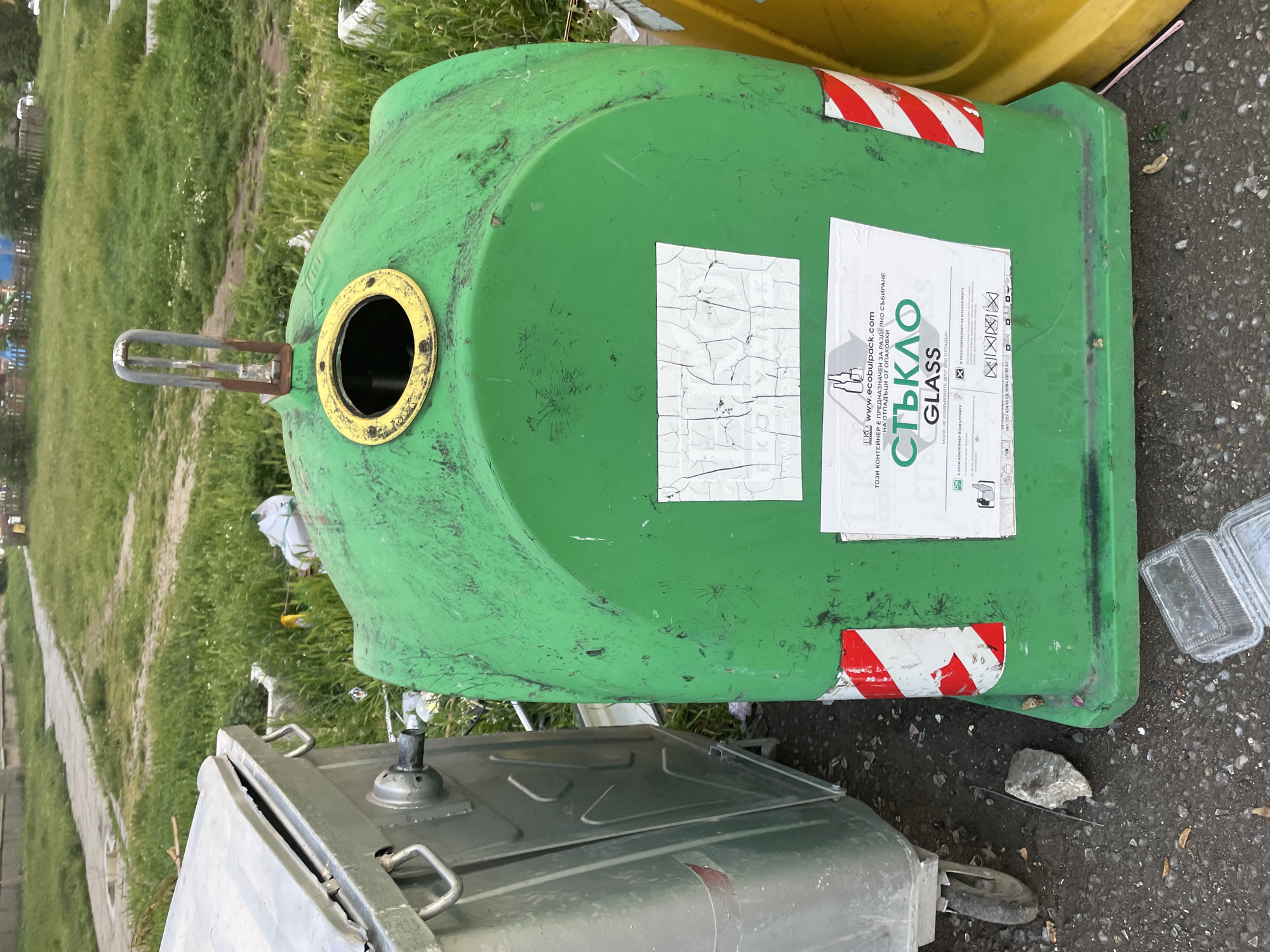 A green recycling bin labeled "GLASS" in Bulgarian and English sits outdoors. The bin is cylindrical with a rounded top, featuring a yellow-rimmed opening and a metal handle. Red and white striped tape is visible on the bin. The bin has stickers, some damaged, with recycling instructions. It is surrounded by grass, a metal bin, and paved asphalt. The location is the city of Sofia, Bulgaria.