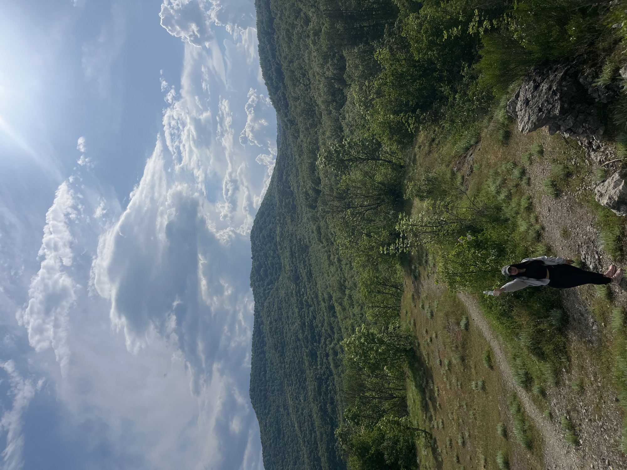 The image shows a person standing on a dirt path in a grassy area in Kamchia, Bulgaria. The person is wearing dark clothing with a light-colored jacket, a hat and holding a plastic bottle. Behind the path is a steep hill covered in dense green forest. Above and to the left, there are puffy white clouds in a light blue sky, with bright sunlight shining from the top left. The image appears to have been rotated to the right.