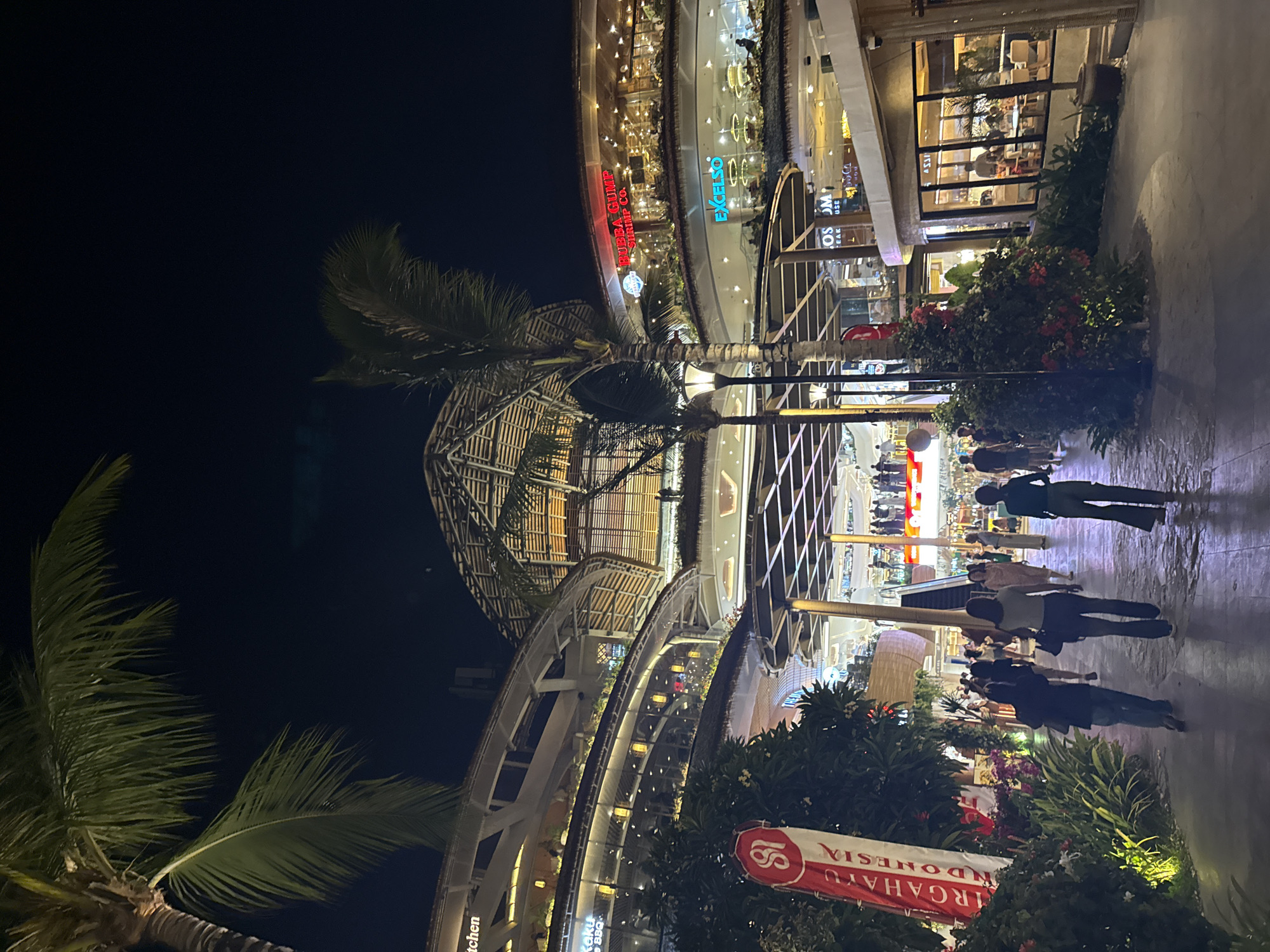 The image depicts a nighttime scene at a large, multi-level open-air shopping and dining complex in Denpasar, Indonesia. The architecture features a distinctive curved, multi-tiered design with exposed bamboo-like roofing structures and extensive use of glass. Palm trees are prominently visible, with their fronds silhouetted against the dark night sky.
The foreground shows a paved walkway where several individuals are walking. On the left side of the walkway, a tall, red and white banner displays the text "S&I MARGAHAYU INDONESIA." Lush green foliage and flowering bushes line the walkway. Lampposts are scattered throughout the scene, providing artificial lighting.
In the background, the various levels of the complex are illuminated. Signs for businesses such as "BUBBA GUMP SHRIMP CO." and "EXCELSO" are visible, indicating a variety of restaurants and shops. People are seen dining and socializing on the upper levels. The overall impression is that of a bustling commercial and entertainment hub in Denpasar.