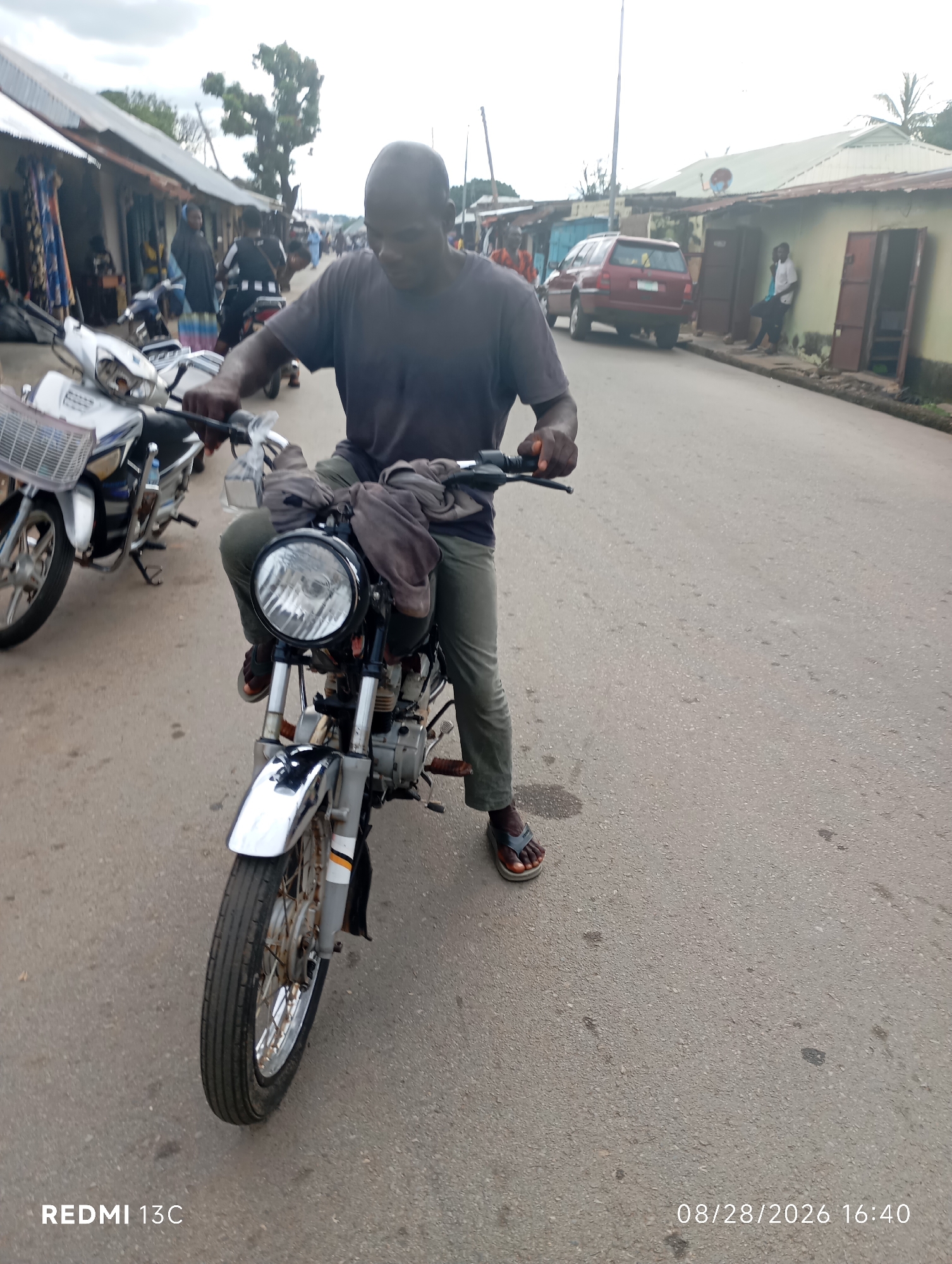A man sits on a motorcycle on a paved road in Shendam, Nigeria. The man is wearing a dark t-shirt and light green pants. He is looking down at the motorcycle's handlebars. The motorcycle is a silver and black model with a basket on the front. In the background, there are buildings and other motorcycles. The sky is overcast. The timestamp indicates the image was taken on August 28, 2026, at 16:40.