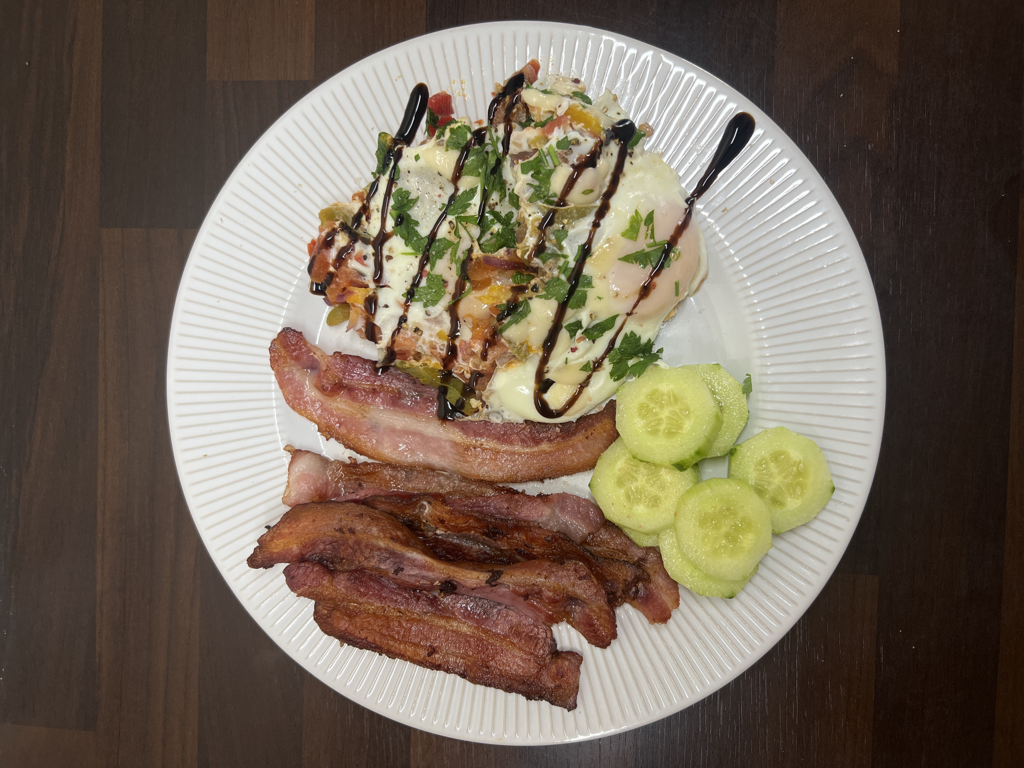 A white plate containing a breakfast of fried eggs, bacon, and sliced cucumbers is displayed on a dark wood table. The fried eggs are topped with chopped vegetables and herbs, and drizzled with a dark sauce. The bacon is cooked to a crisp and arranged to the left of the eggs. Several slices of cucumber are placed to the bottom left of the plate. The table surface suggests a domestic setting. There are no individuals present. The image provides a clear, overhead view of the meal.