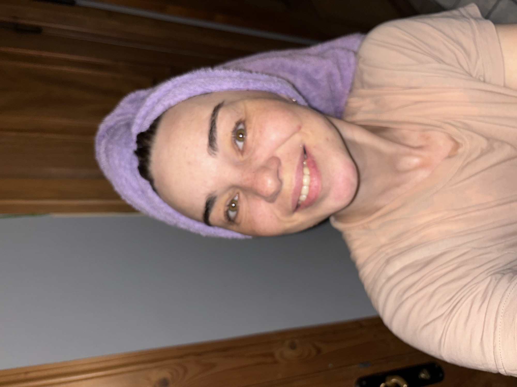 A woman with fair skin, brown hair covered by a lavender-colored towel, and hazel eyes is smiling. She is wearing a light peach-colored, long-sleeved shirt. The background consists of a light gray wall in the middle and a wooden surface with black and gold hardware in the lower right. Above the woman, there is a wooden panel with a textured surface. The location and city are unknown.