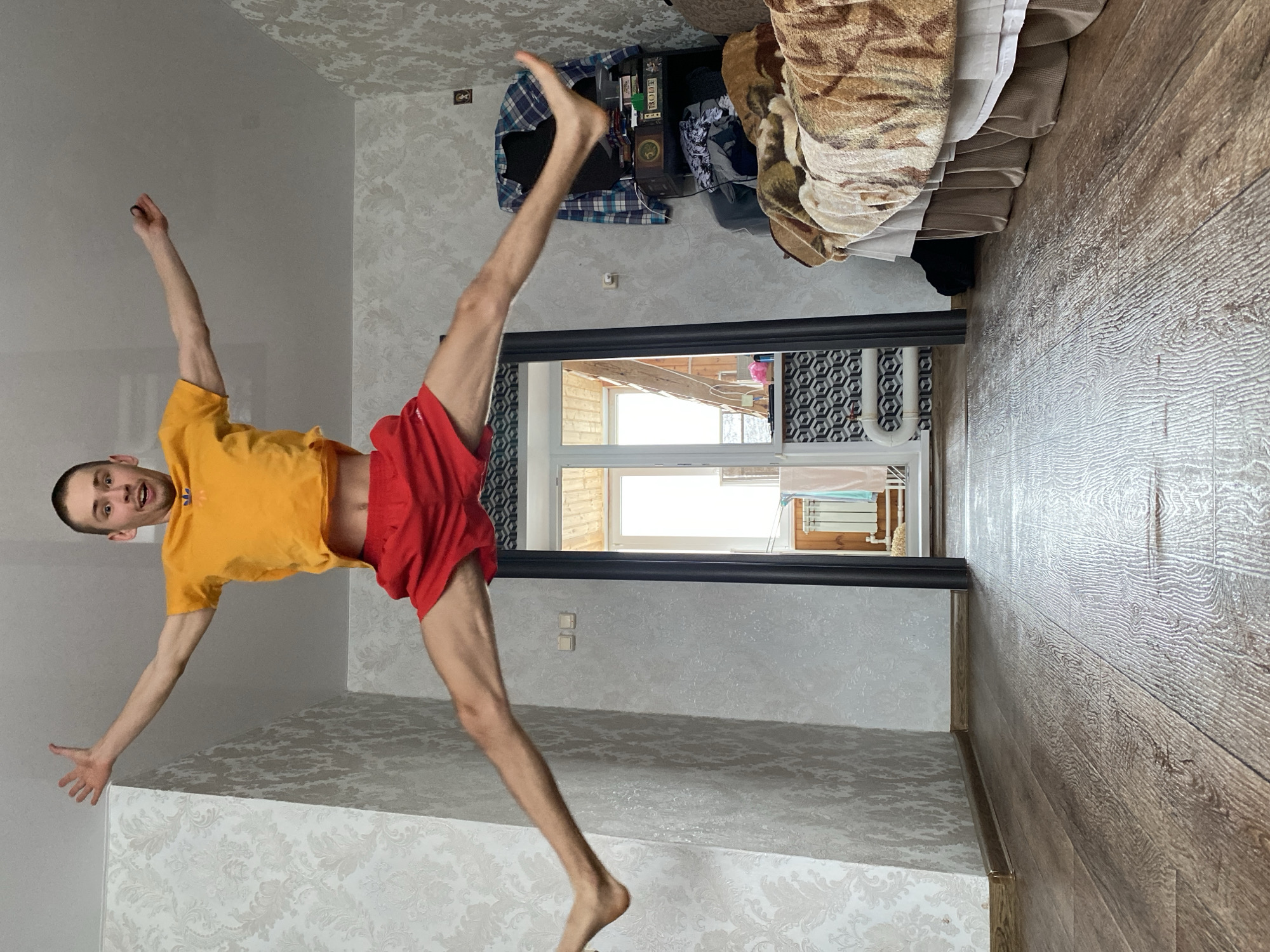 An energetic, full-body shot captures a young man in mid-air within an indoor setting in Ulyanovsk, Russia. The man, with short dark hair and a fair complexion, wears a bright yellow t-shirt and red shorts, and is barefoot. He is caught in a dynamic jump or acrobatic pose, with his arms and legs spread widely. His mouth is open, conveying exertion or excitement. His right arm and right leg are extended upwards, while his left arm is extended outwards, slightly bent at the elbow with an open hand, and his left leg is bent at the knee, pointing downwards.
The room features light-colored walls adorned with a subtle, ornate damask wallpaper pattern, and a light brown wooden floor with a distinct grain. In the background, a dark-framed doorway opens to what appears to be a balcony or outdoor area, revealing a window and an external view of light-colored wooden structures. Inside the room, to the right, a bed or sofa with patterned covers (brown and beige, possibly animal print) is visible, alongside a dark piece of furniture (possibly a dresser or shelf) with clothes piled on it. A box with the text "BOTT" is visible on this furniture. The bright lighting suggests it is daytime with clear weather outside. Wall-mounted light switch or outlet plates are also noticeable near the doorway.