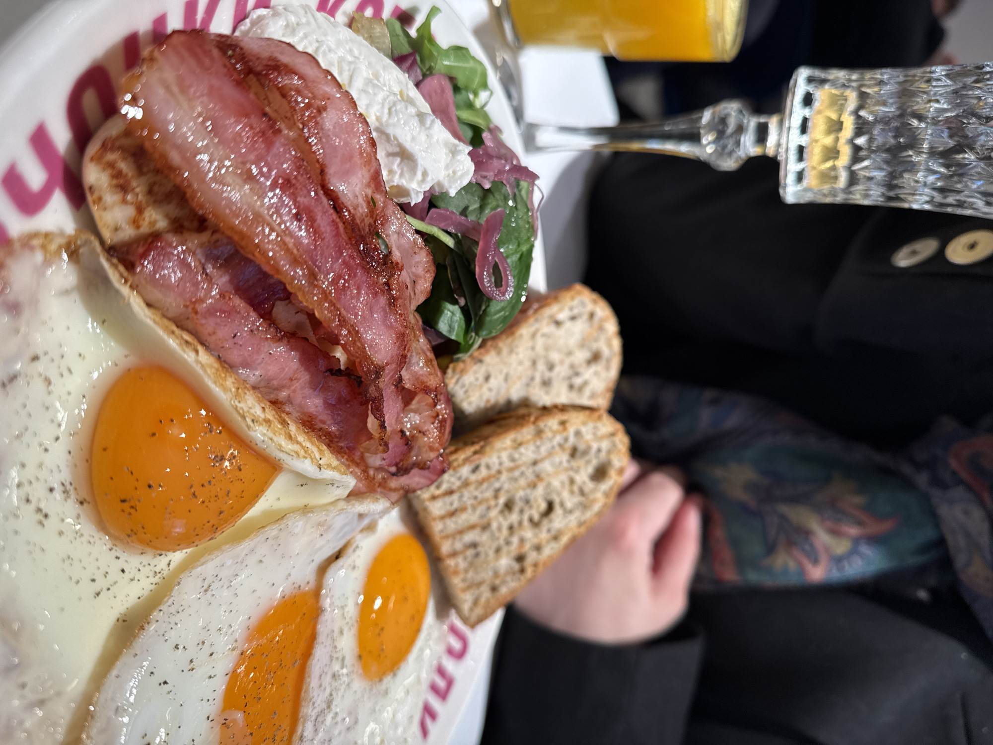 A plate of food is positioned prominently in the image. It contains two fried eggs, several strips of bacon, a dollop of a white creamy substance atop a small salad, and two pieces of toast. A glass of orange juice stands nearby, with a crystal-cut glass stem. A person is sitting in what appears to be a black jacket, with buttons visible, holding a piece of cloth. The scene is likely indoors. There is not enough information to determine the specific location in Moscow or to confirm any actions, interactions, or further environmental details.