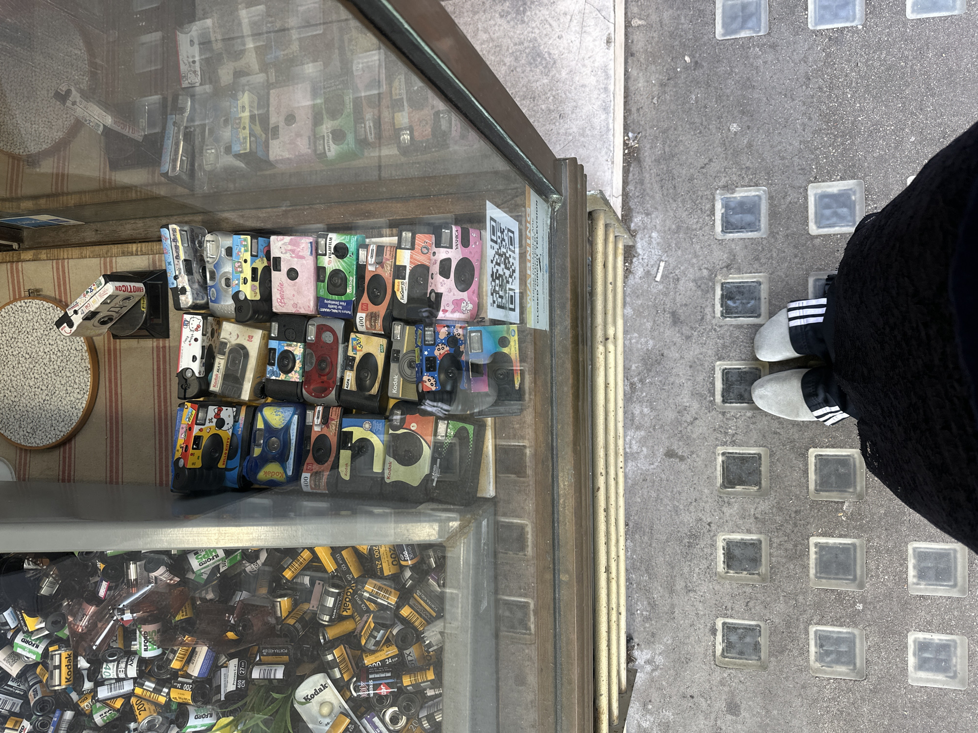 A top-down view captures a shop display case filled with disposable cameras and film rolls. The cameras, arranged on the top shelf, are in various colors and patterns, including a "Hello Kitty" design. Below them, a jumble of Kodak, Ilford, and other film rolls occupies the lower shelf. A sign with a QR code is affixed to the glass case. The display sits on a sidewalk made of square glass blocks set into the concrete. A person's legs, clad in dark clothing and wearing white Adidas shoes with black stripes, are visible near the case. The photo was taken in the city of London, United Kingdom.