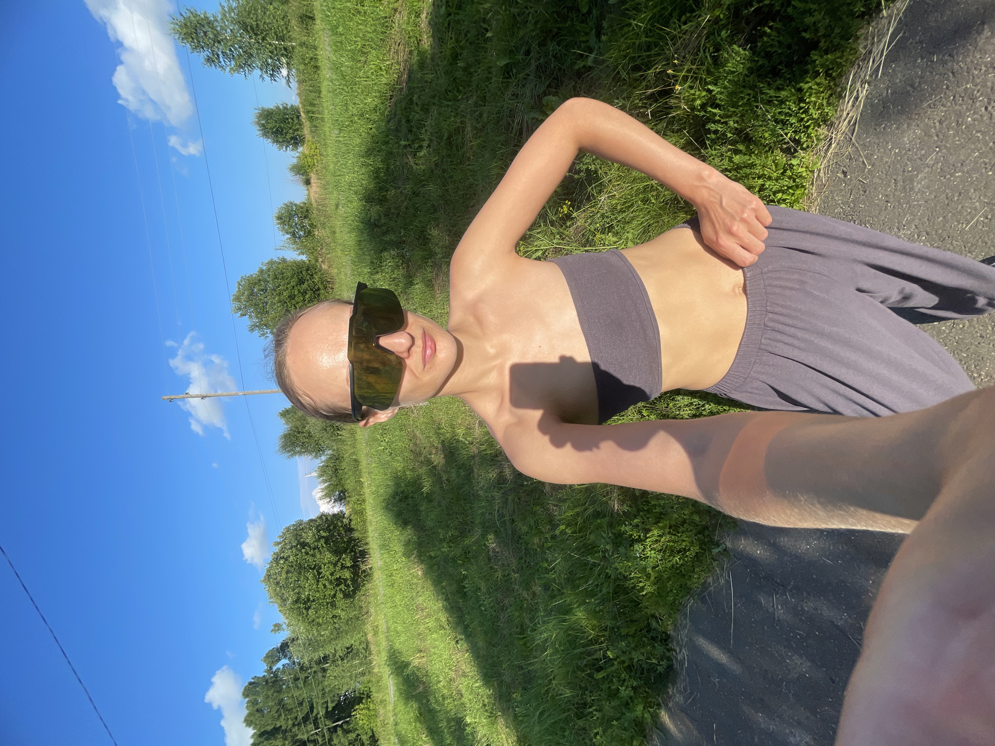 The image depicts a person standing outdoors in a rural setting, likely Podshivalovo, Russia. The subject, a woman, is wearing a grey sports bra and loose-fitting grey pants, along with large, dark sunglasses. She is posing for a selfie, with her right arm extended and her hand resting on her pants near her waist. Her left arm is bent at the elbow, with her hand resting on her hip. The scene is a grassy field with trees in the background. The sky is a clear, bright blue with a few scattered white clouds, suggesting a sunny day. The sun is casting distinct shadows on the grass, indicating it is likely daytime, possibly late morning or early afternoon. The overall impression is one of casual recreation or exercise in a natural environment. There is no visible text in the image.
