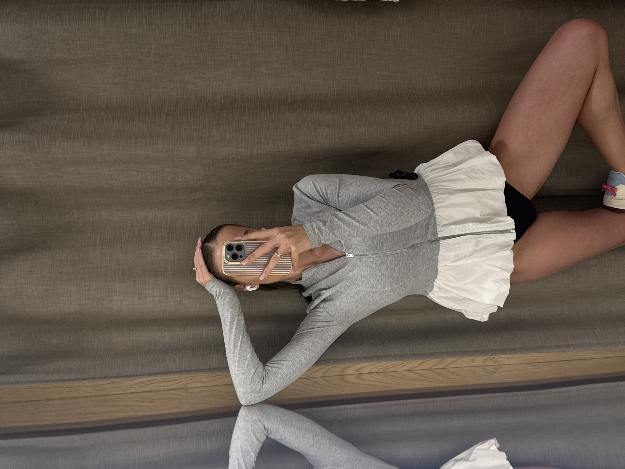 A woman lies on a couch, taking a selfie in a mirror. She is wearing a grey zip-up jacket over a white ruffled skirt and black shorts. Her hair is dark brown and pulled back. She has on white wireless earbuds and rings on her fingers. Her right hand is holding up her phone, which has a gold case. Her left arm is bent and her hand is resting on her head. Her legs are crossed and she is wearing white sneakers with pink laces and bows. The couch appears to be made of a dark grey, textured fabric. A wooden shelf is visible below the couch. The lighting suggests it is daytime, but there are no windows or outdoor elements to confirm. The mood seems relaxed and casual.