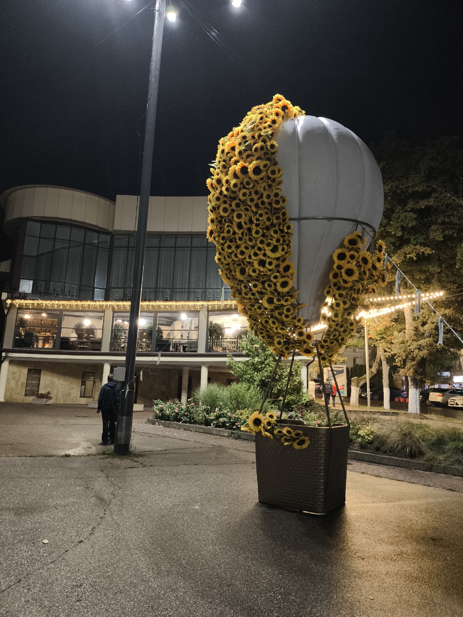 This image depicts a nighttime scene in Yessentuki, Russia, featuring a large, illuminated hot air balloon installation adorned with numerous sunflowers. The balloon, predominantly white with a metallic sheen, is covered on its upper half and sides with bright yellow sunflowers, some of which are lit by spotlights. The installation is anchored by a large, dark woven basket at its base. In the background, a modern building with large glass windows and a curved facade is visible. String lights are draped across the facade and illuminated, casting a warm glow. A single person, wearing a dark jacket and pants, stands near a tall lamppost in the foreground, facing away from the camera. The ground is wet and reflective, suggesting recent rain. Trees and foliage are present on the right side of the image, along with parked cars on a street in the distance. The overall atmosphere is festive and visually striking, with the bright sunflowers contrasting against the dark night sky and the illuminated building.