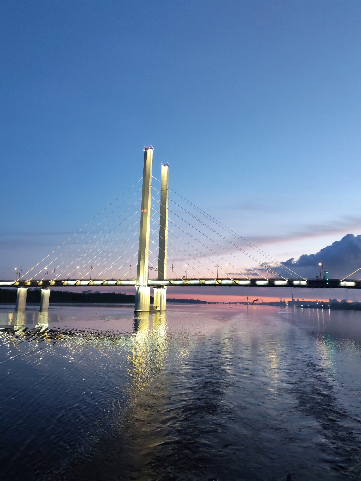 The image shows the October Bridge in Cherepovets, Russia at dusk. The bridge, a cable-stayed structure, is the primary subject, with two tall, illuminated towers supporting the roadway via numerous cables. The bridge and its reflection are visible in the water below. The sky is a gradient of blue to pink, with some clouds visible on the horizon. Distant industrial buildings can be seen along the horizon. Lights illuminate the bridge and reflect off the water's surface, creating a shimmering effect.