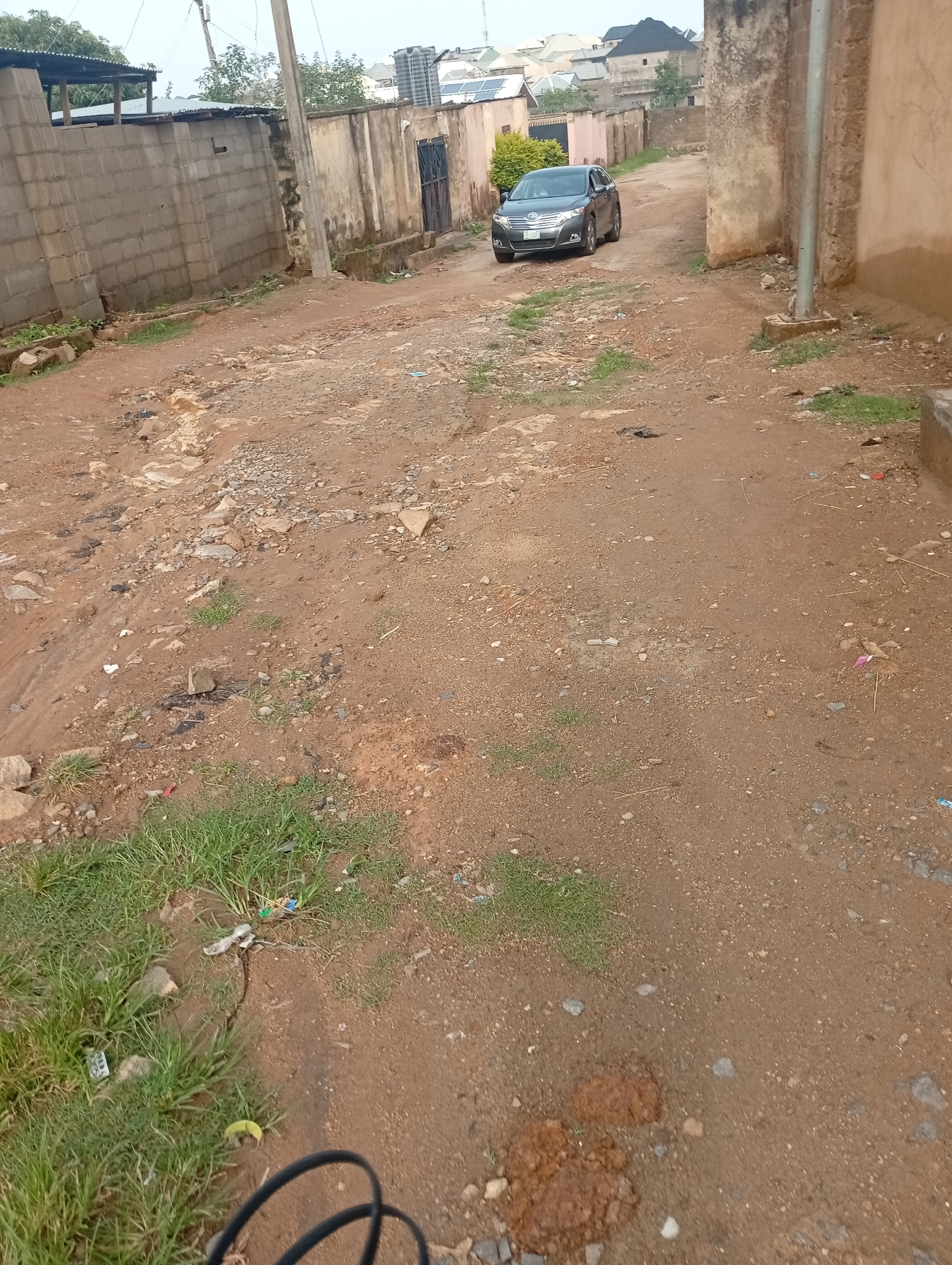This is a ground-level, slightly angled shot looking down a dusty, unpaved road in Rafin Par, Nigeria. The road is lined with buildings on either side, constructed with a mix of concrete blocks and rendered walls. On the left, a high, grey concrete block wall runs along the side of the road, partially obscured by some vegetation. Further down the road on the left, there are more buildings, some with corrugated metal roofs. On the right, a textured, weathered, tan-colored wall dominates the foreground, with a simple metal utility pole in front of it.
A dark gray sedan is parked on the right side of the road, facing away from the camera. The car has a tinted windshield and a visible license plate. The road surface is predominantly red-brown earth mixed with gravel and scattered small stones. Patches of green grass are growing sporadically along the sides of the road, particularly in the foreground on the left, where some litter is also visible. In the background, more buildings with various roof types, including solar panels, are visible, suggesting a residential area. The sky is overcast, contributing to a diffused light.