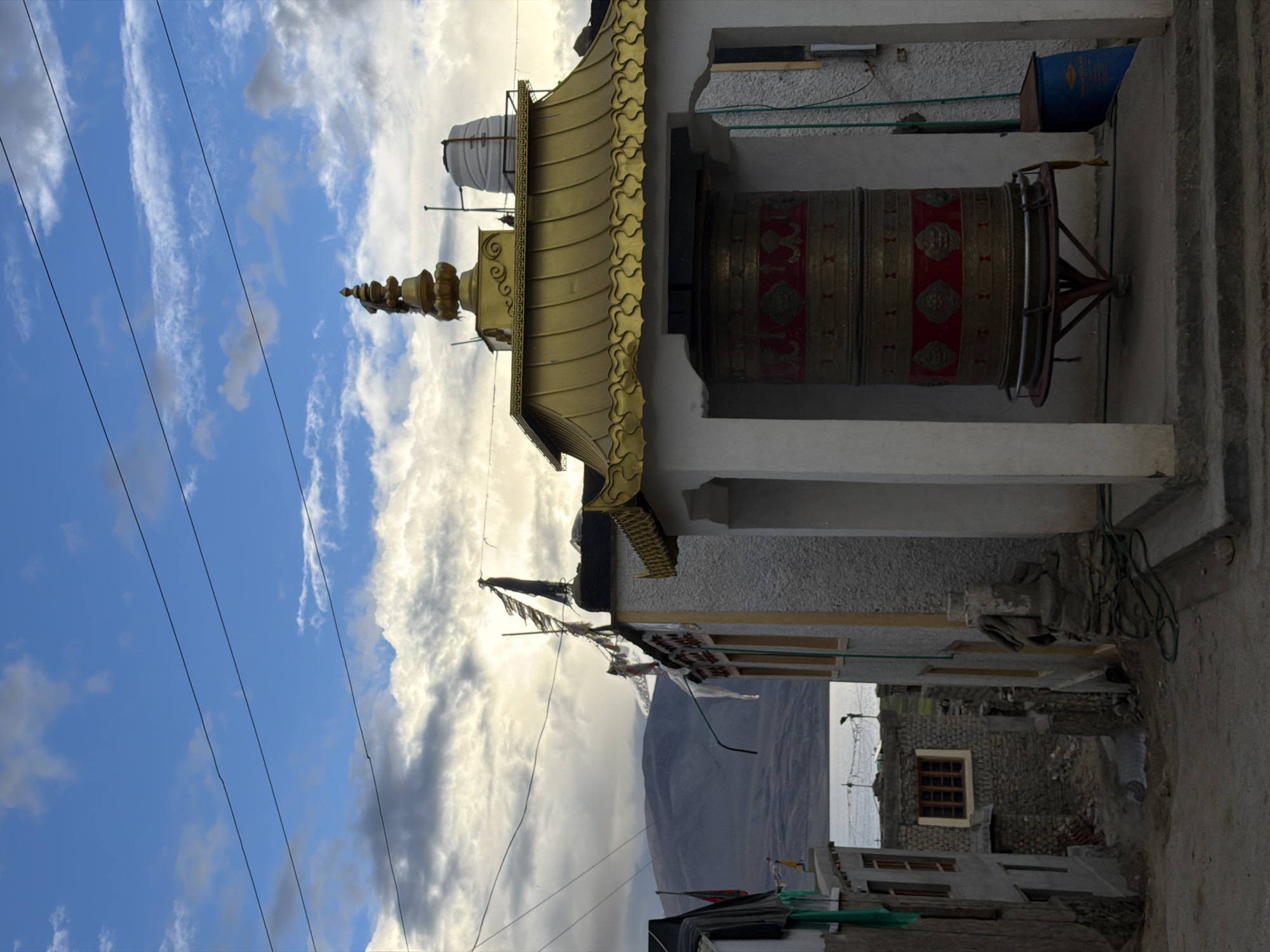 The image shows a prayer wheel in the village of Karzok, India, a village located on the shore of Tso Moriri Lake in Ladakh. The prayer wheel is large, cylindrical, and ornately decorated with red and gold patterns. It is housed in an open structure with white pillars and a golden, tiered roof. The prayer wheel is mounted on a dark red stand. In the background, there are several buildings with white and textured walls, some with prayer flags strung across them. Mountains and a body of water are visible beyond the village. The sky is a mix of blue with white and grey clouds. The overall scene depicts a cultural and religious element of the village.