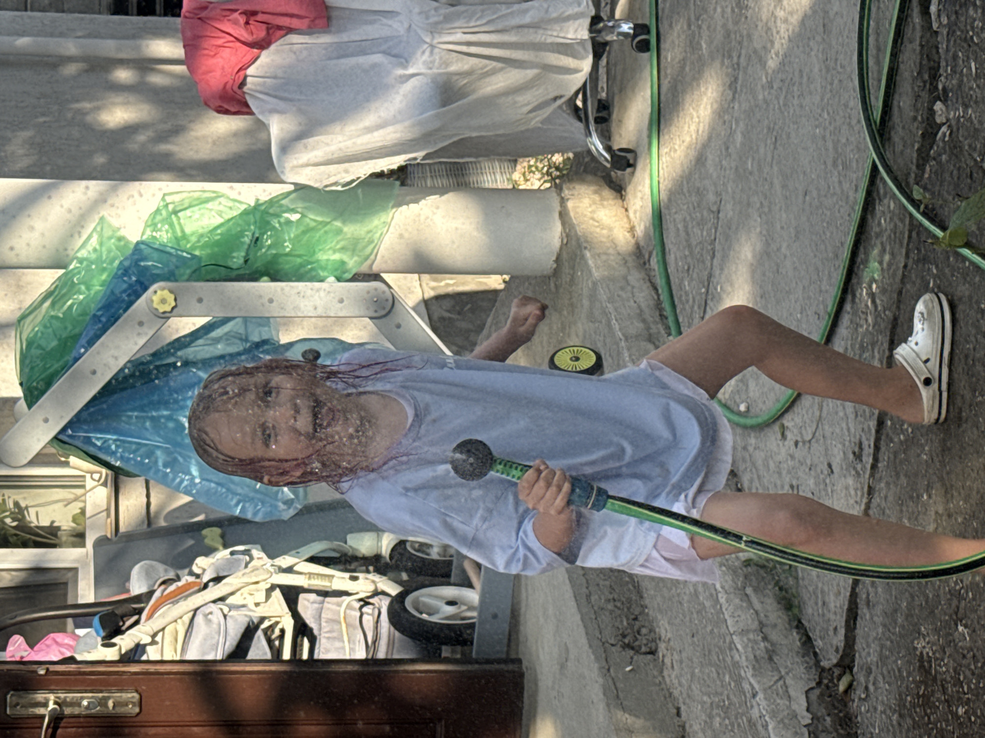 A young, fair-skinned child with reddish-pink hair is captured mid-action in an outdoor concrete area in Unknown, Unknown. The child, appearing to be female, wears a light blue oversized t-shirt, white shorts, and a single white Croc-style shoe on the right foot. Her face is extensively covered in water droplets, and she displays a wide, open-mouthed smile, looking directly towards the viewer. She holds a green garden hose with a black spray nozzle in both hands, actively directing water outwards.
The immediate ground is concrete, wet and showing signs of wear and cracking. To the left, a brown wooden door frame with a brass handle is visible, partially obscuring a stack of light-colored, possibly fabric-covered, wheeled items or strollers. In the background, a light-colored building wall features a window with a visible plant. A metallic structure, possibly a ladder or scaffold part, stands upright, adorned with draped blue and green plastic sheeting. Further to the right, a white sheet covers an object with chrome legs and small wheels, likely a chair or cart, topped with a folded piece of red fabric. Strong sunlight illuminates the scene, casting distinct shadows from the structures and the child.