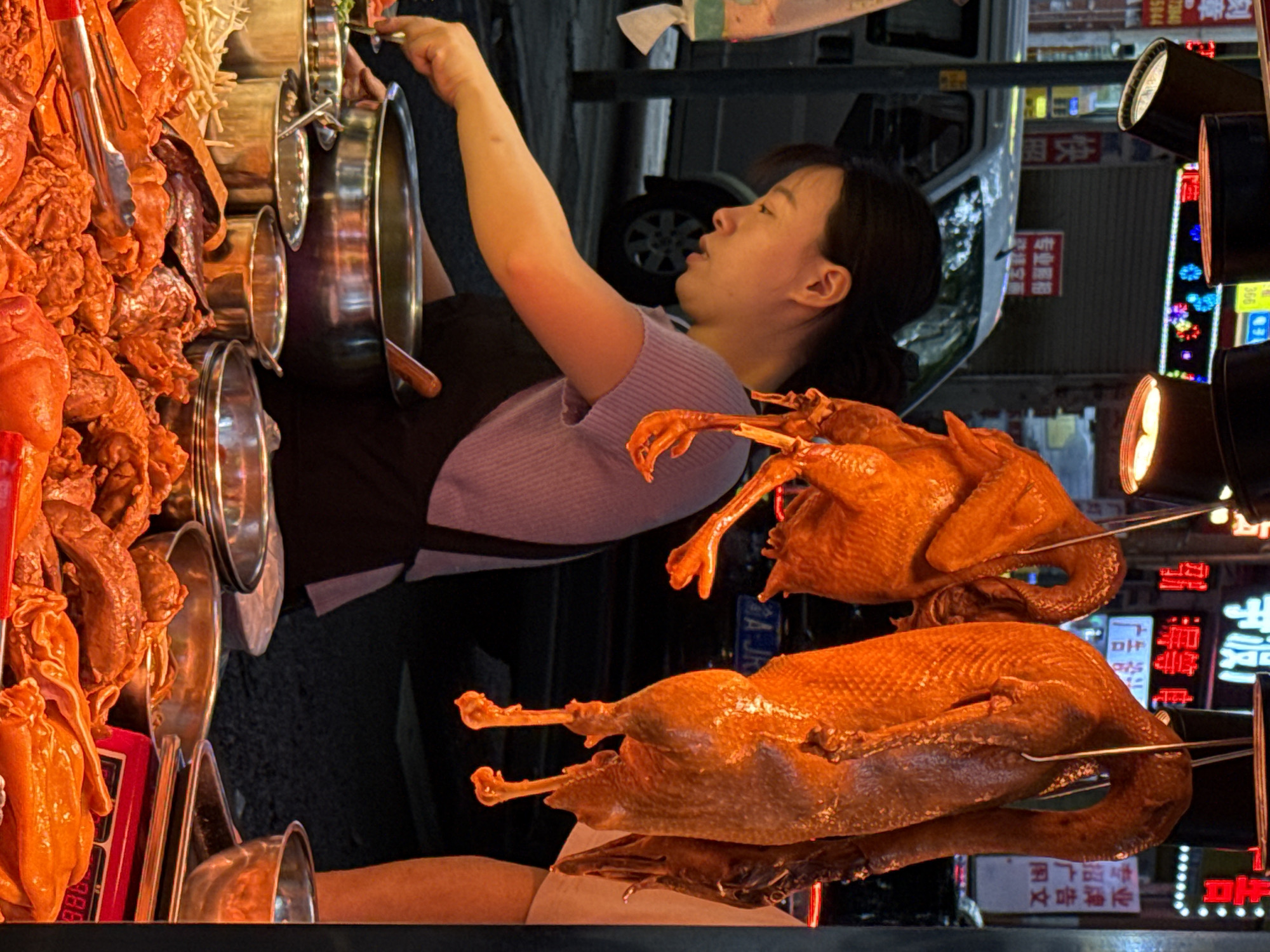 Here is a detailed description of the image:
The image shows a food stall in Nan'an District, China. An Asian woman is standing behind the stall, tending to some food in metal bowls. She is wearing a purple top and a dark apron.
On the left side of the frame, there is an assortment of cooked meats displayed, likely for sale. To the right of the woman, two roasted ducks are skewered and hanging in the air, ready to be sold.
In the background, there is a blurred view of a street with a white van parked. Streetlights and neon signs are visible, indicating the setting is likely at night. Some of the neon signs have Chinese characters.