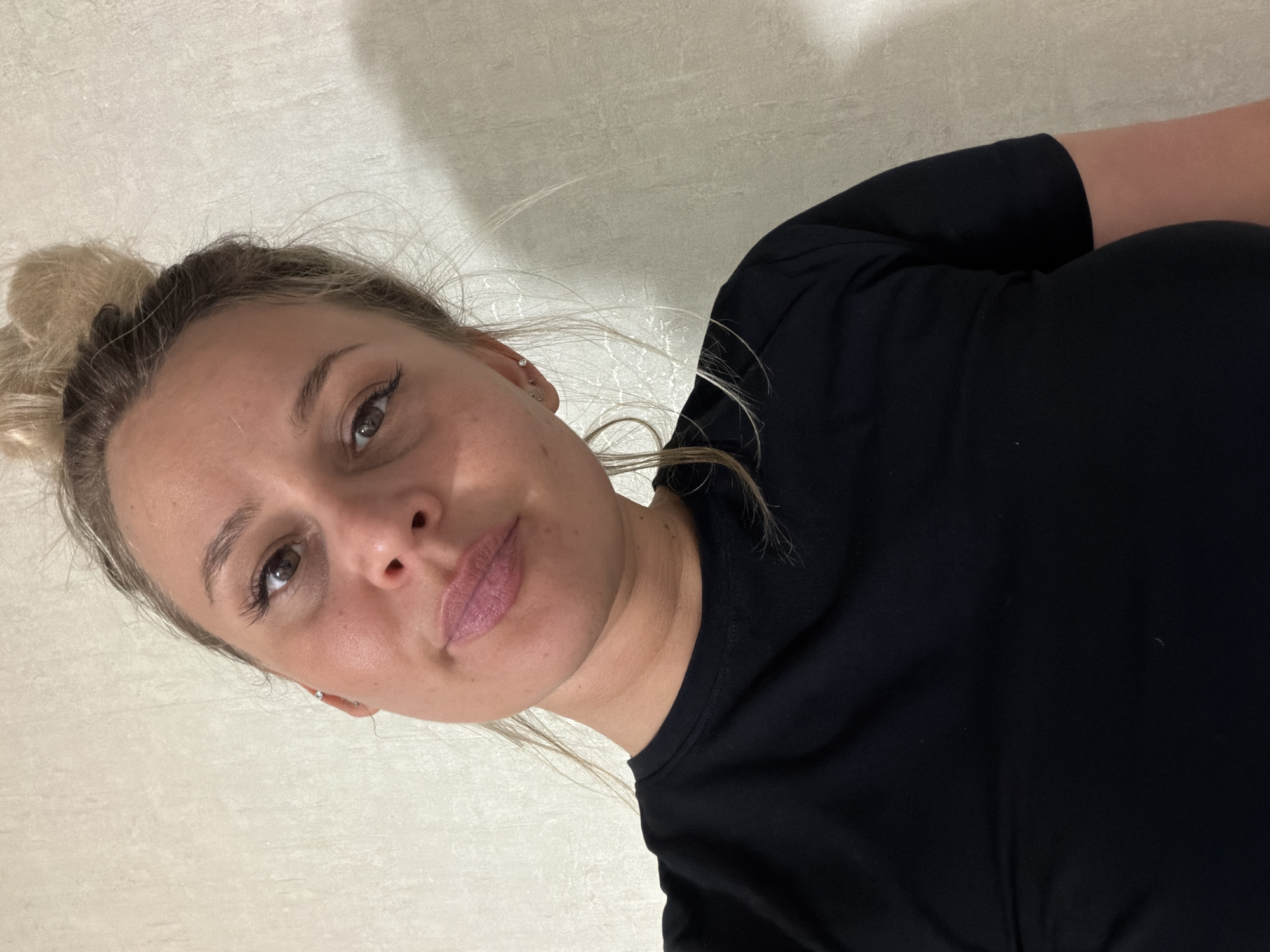 A medium close-up angled shot shows a woman with blonde hair pulled up in a messy bun. She has fair skin, brown eyes, and is wearing pink lipstick. A small earring is visible in her right ear. She is wearing a black t-shirt. The background is a light beige or gray textured wall. The location in the city of Sarapul, Russia is not identifiable in the provided image.
